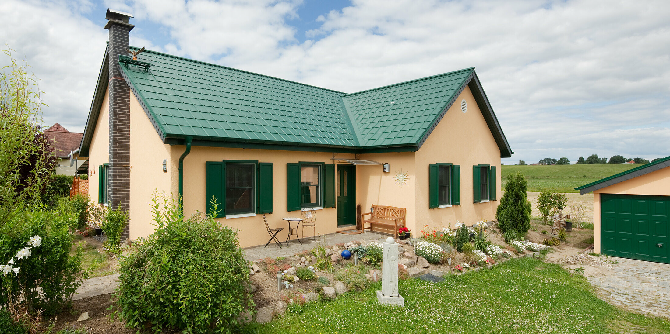 Die reizvolle Gesamtansicht des Einfamilienhauses in Friedrichswalde zeigt das langlebige Aluminiumdach in Moosgrün, das sowohl das Wohnhaus als auch die Garage ziert. Der grüne Farbton der PREFA Dachplatten fügt sich harmonisch in die natürliche Umgebung ein und unterstreicht die schlichte Architektur des Hauses. Das erweiterte Satteldach in L-Form liegt vor einem Feld. Das schmucke Häuschen hat passende grüne Fensterläden und einen hübschen kleinen Vorgarten.