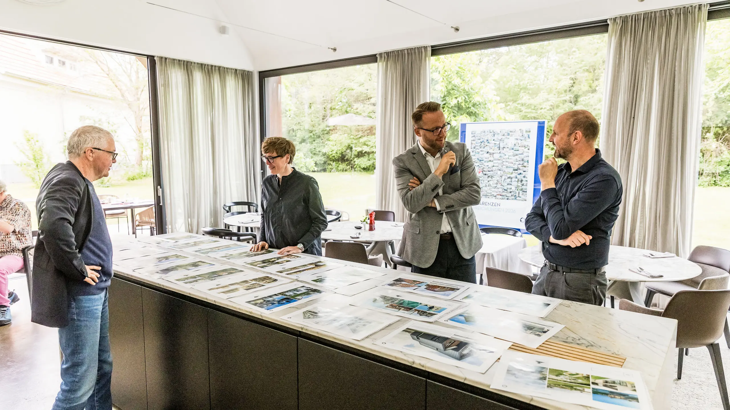 Die finale Auswahl der PREFARENZEN Architekturhighlights für 2026 liegt auf großen Bildern vor Teilen der hochkarätig besetzten Jury. Die Experten prüfen die eingereichten Architekturprojekte sorgfältig auf Innovationskraft, gestalterische Qualität und den Einsatz von PREFA-Materialien. Die Sitzung in der lichtdurchfluteten Winzerresidenz im burgenländischen Seewinkel bildet den Abschluss des intensiven Juryprozesses für das PREFARENZEN-Architekturbuch und den Kalender 2026.