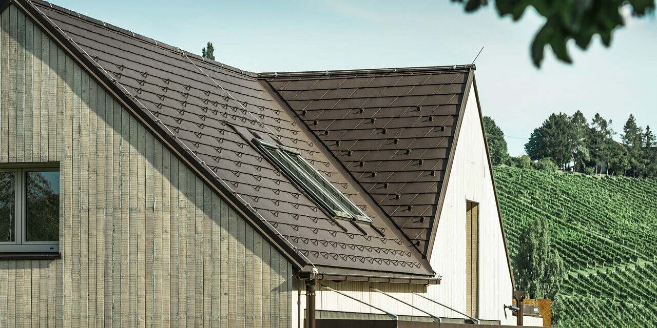 Das Einfamilienhaus mit zwei Satteldächern wurde mit der PREFA Dachschindel in Nussbraun eingedeckt. Die Dachentwässerung erfolgt über die PREFA Kastenrinne. Die Fassade ist mit verwittertem Holz verkleidet.