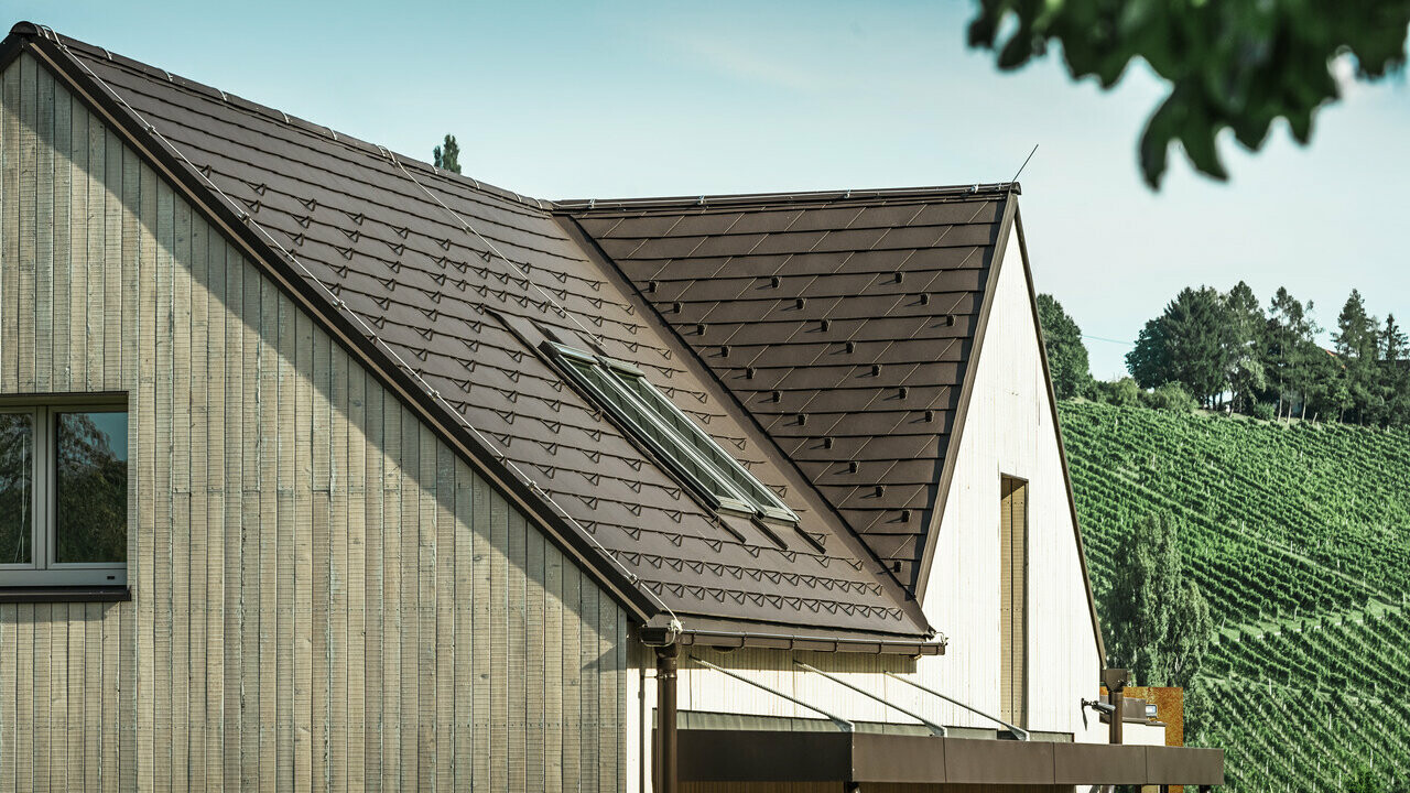 Das Einfamilienhaus mit zwei Satteldächern wurde mit der PREFA Dachschindel in Nussbraun eingedeckt. Die Dachentwässerung erfolgt über die PREFA Kastenrinne. Die Fassade ist mit verwittertem Holz verkleidet.