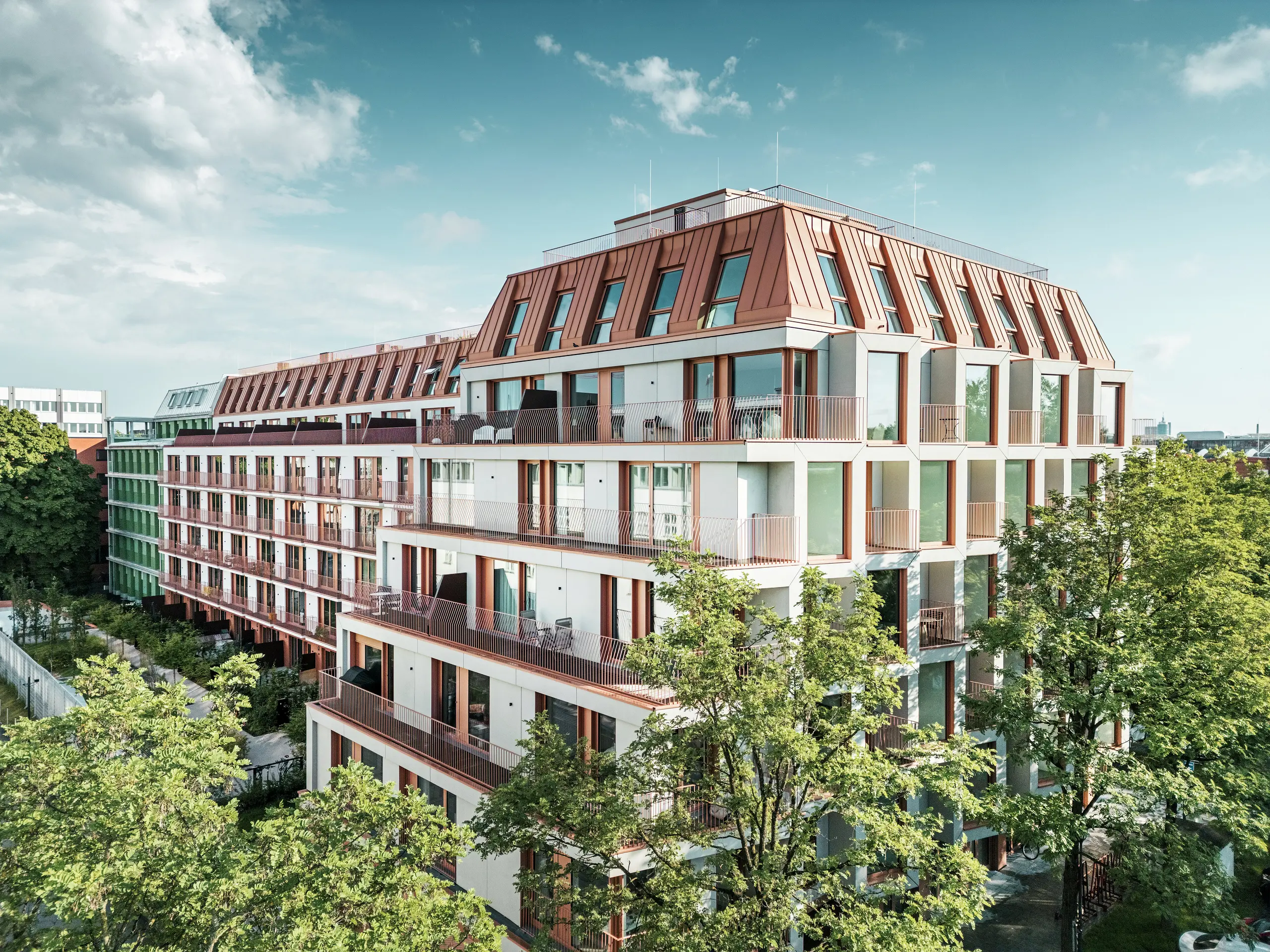 Der Blick auf das urbane Wohnprojekt Van B in München besticht durch seine klare Architektur, die begrünten Außenbereiche und das markante PREFALZ-Aluminiumdach in IGP Orange HDP von PREFA. Das langlebige und pflegeleichte Dachmaterial bildet ein verbindendes Gestaltungselement des Gebäudes und bietet eine dauerhafte, ästhetische Lösung im urbanen Raum. Großflächige Glasfronten, Balkone und ein durchdachter Grundriss ermöglichen modernen Wohnkomfort mit hoher Designqualität.