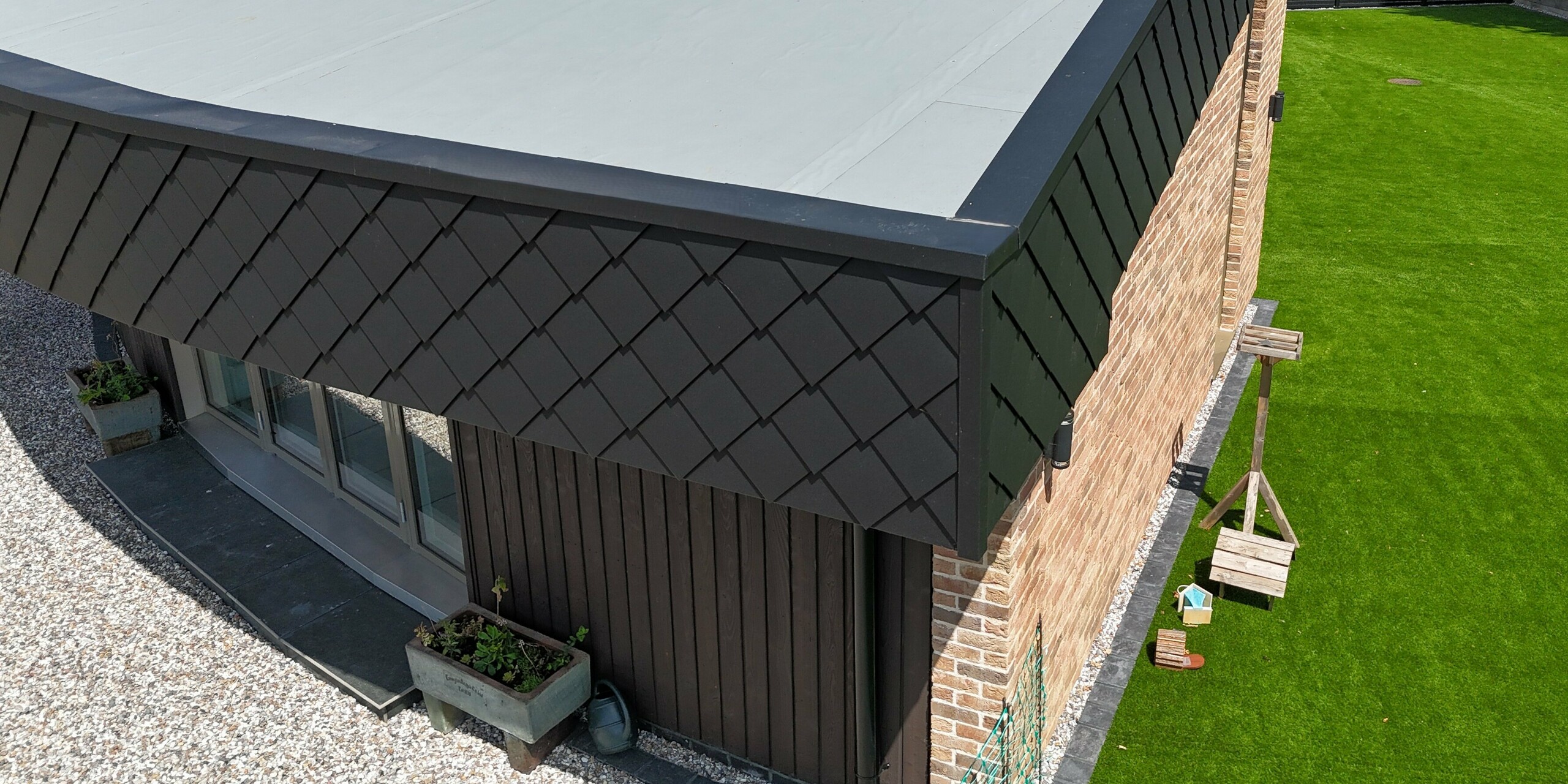 Detailansicht eines modernen Eco Homes mit markanter Gebäuderundung, die aus einer Kombination von warmem Klinker und der eleganten PREFA Wandraute 20 × 20 in Anthrazit besteht. Die Aluminium-Elemente setzen einen starken Kontrast und verleihen dem Haus eine dynamische, hochwertige Optik. Dank der robusten PREFA-Produkte ist die Fassade extrem langlebig, witterungsbeständig und pflegeleicht – ideal für nachhaltige Architektur. Im Vordergrund sind die klaren Linien und die präzise Verarbeitung erkennbar, die das Zusammenspiel von Design und Funktionalität perfekt unterstreichen.