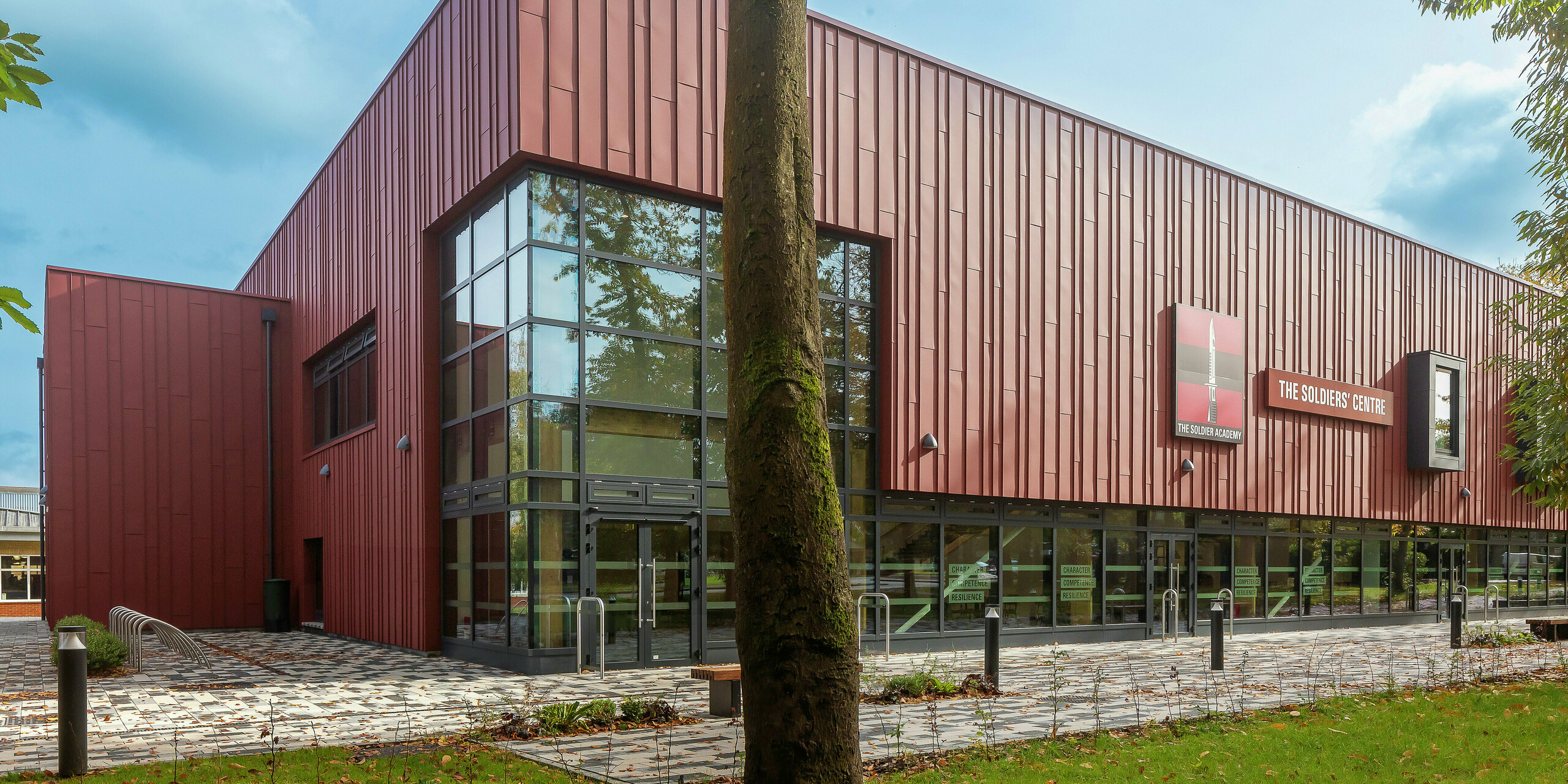 Innovative Fassadengestaltung mit PREFALZ: Das Soldiers' Centre in Pirbright beeindruckt mit einer modernen Stehfalzverkleidung aus PREFALZ in P.10 Oxydrot. Großzügige Glasflächen sorgen für eine helle, einladende Atmosphäre, Fensterrahmen in P.10 Anthrazit setzen stilvolle Akzente. Nachhaltigkeit, Langlebigkeit und Design vereint - hochwertige PREFA Aluminiumprodukte für zukunftsweisende Architektur.