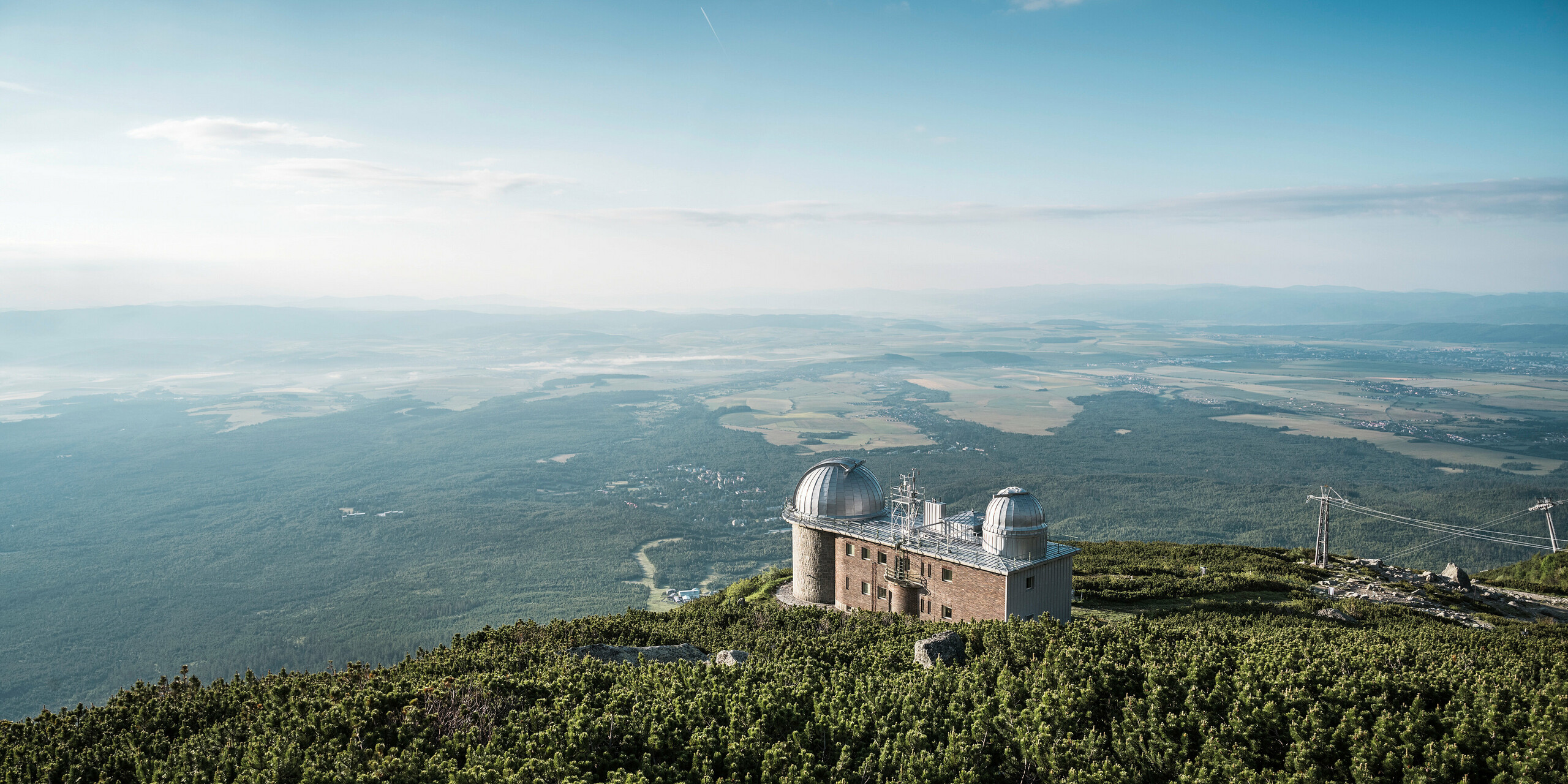 Atemberaubende Fernsicht auf die Sternwarte Skalnaty Pleso, gelegen in 1.783 m Höhe über dem Meeresspiegel, umgeben von der ruhigen Pracht der Hohen Tatra in der Slowakei. Die beiden charakteristischen Kuppeln, bedeckt mit glänzendem PREFALZ in Silbermetallic, reflektieren die Weite des Himmels und harmonieren mit der natürlichen Umgebung. Das Observatorium steht nicht nur für astronomische Exzellenz, sondern auch für die Beständigkeit und ökologische Stärke von PREFA, dessen Aluminiumprodukte selbst in dieser entlegenen Höhe Schutz und Beständigkeit bieten, was durch eine umfassende 40-jährige Garantie bekräftigt wird.