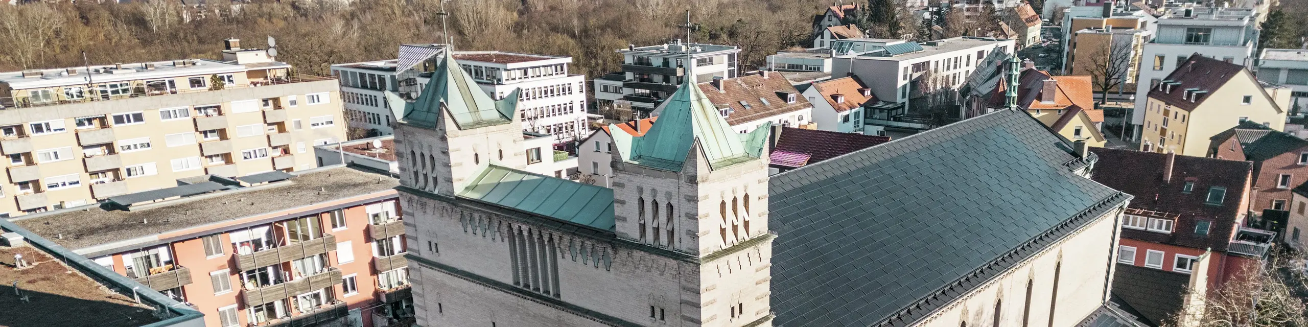 Vorderseite der St. Karl Borromäus Kirche in Nürnberg: Das Bild zeigt das beeindruckende Dach der denkmalgeschützten Kirche, das mit schwarzen PREFA-Solardachplatten gedeckt ist. Die Solarmodule fügen sich harmonisch in die historische Architektur der Kirche ein und stellen gleichzeitig eine moderne Lösung zur nachhaltigen Energiegewinnung dar. Im Hintergrund sind moderne Gebäude und die grünen Hügel Nürnbergs zu sehen, was den gelungenen Übergang zwischen traditioneller Kirchenarchitektur und zeitgemäßer Energieversorgung verdeutlicht. Das Aluminiumdach der Kirche wird durch die stilvolle PREFA Dachplatte R.16 vervollständigt.