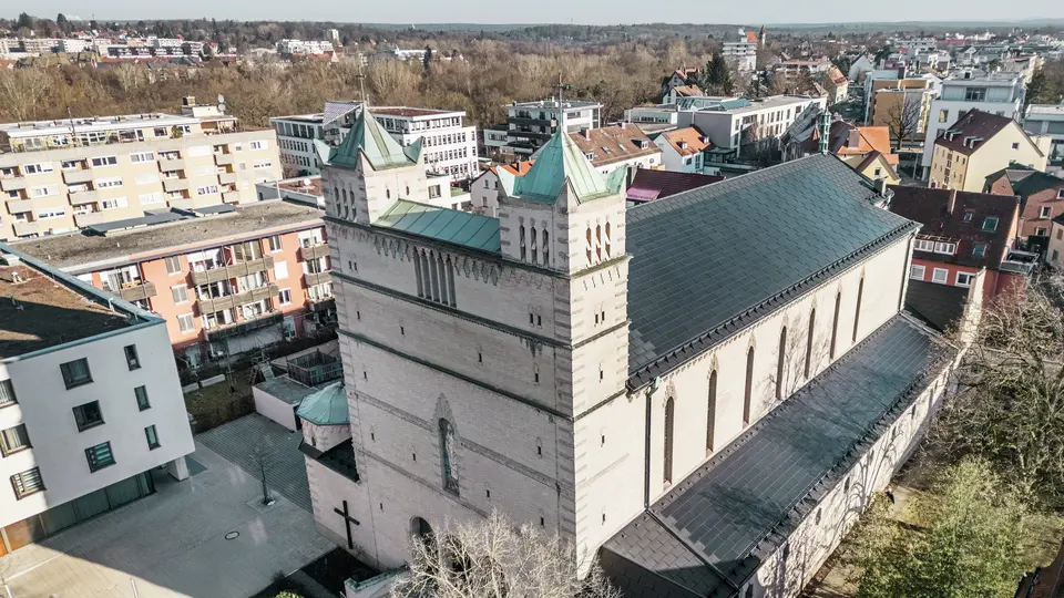 Vorderseite der St. Karl Borromäus Kirche in Nürnberg: Das Bild zeigt das beeindruckende Dach der denkmalgeschützten Kirche, das mit schwarzen PREFA-Solardachplatten gedeckt ist. Die Solarmodule fügen sich harmonisch in die historische Architektur der Kirche ein und stellen gleichzeitig eine moderne Lösung zur nachhaltigen Energiegewinnung dar. Im Hintergrund sind moderne Gebäude und die grünen Hügel Nürnbergs zu sehen, was den gelungenen Übergang zwischen traditioneller Kirchenarchitektur und zeitgemäßer Energieversorgung verdeutlicht. Das Aluminiumdach der Kirche wird durch die stilvolle PREFA Dachplatte R.16 vervollständigt.