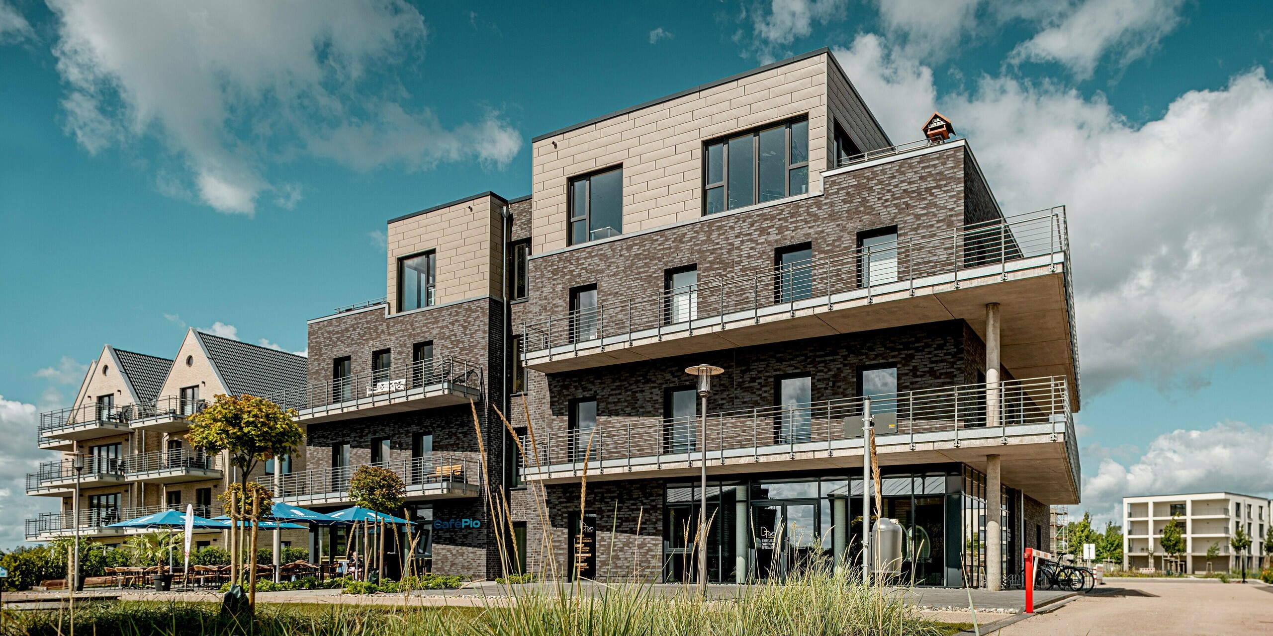 Das Foto zeigt ein Gebäude der Wohnanlage „Hafen zur Freiheit“ mit einer kraftvollen Kombination aus dunkler Klinkerfassade und dem darüberliegenden Staffelgeschoss aus PREFA Siding.X in edlem Bronze. Die strukturierten Aluminiumoberflächen verleihen dem oberen Bereich eine moderne, hochwertige Tiefe und setzen einen deutlichen Kontrast zum traditionellen Mauerwerk. Großzügige Balkone, markante Linien und viel Glas betonen die offene Architektur. Die witterungsbeständigen, langlebigen PREFA Paneele sorgen für dauerhaften Schutz und eignen sich ideal für die exponierte Küstenlage.