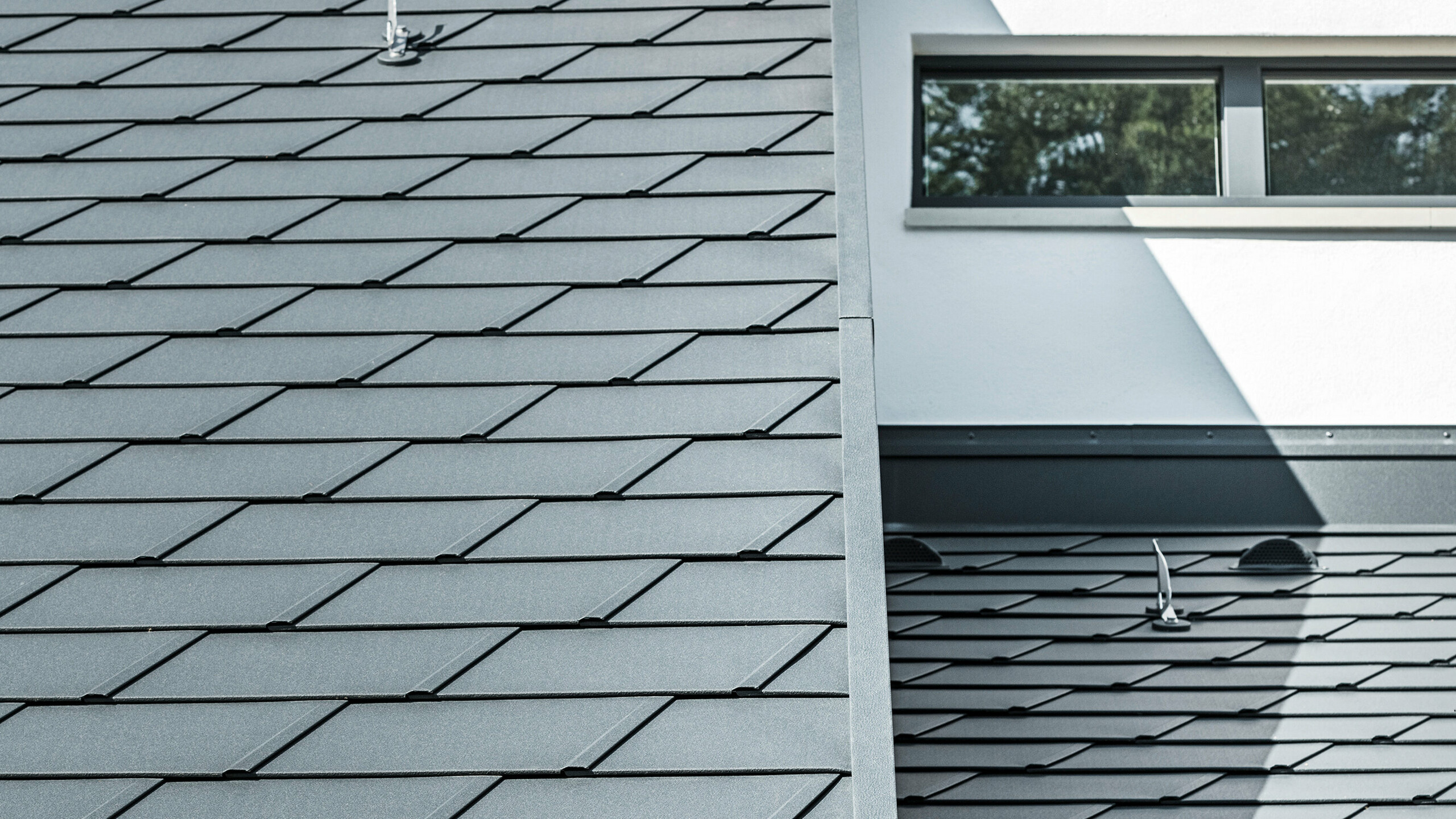 Detailaufnahme der unterschiedlichen Ebenen am Dach eines Einfamilienhauses in Dortmund. Zu sehen sind die PREFA Dachschindel DS.19 und das PREFA Dachentwässerungssystem in der Farbe P.10 Anthrazit sowie die PREFA Sicherheitsdachhaken.