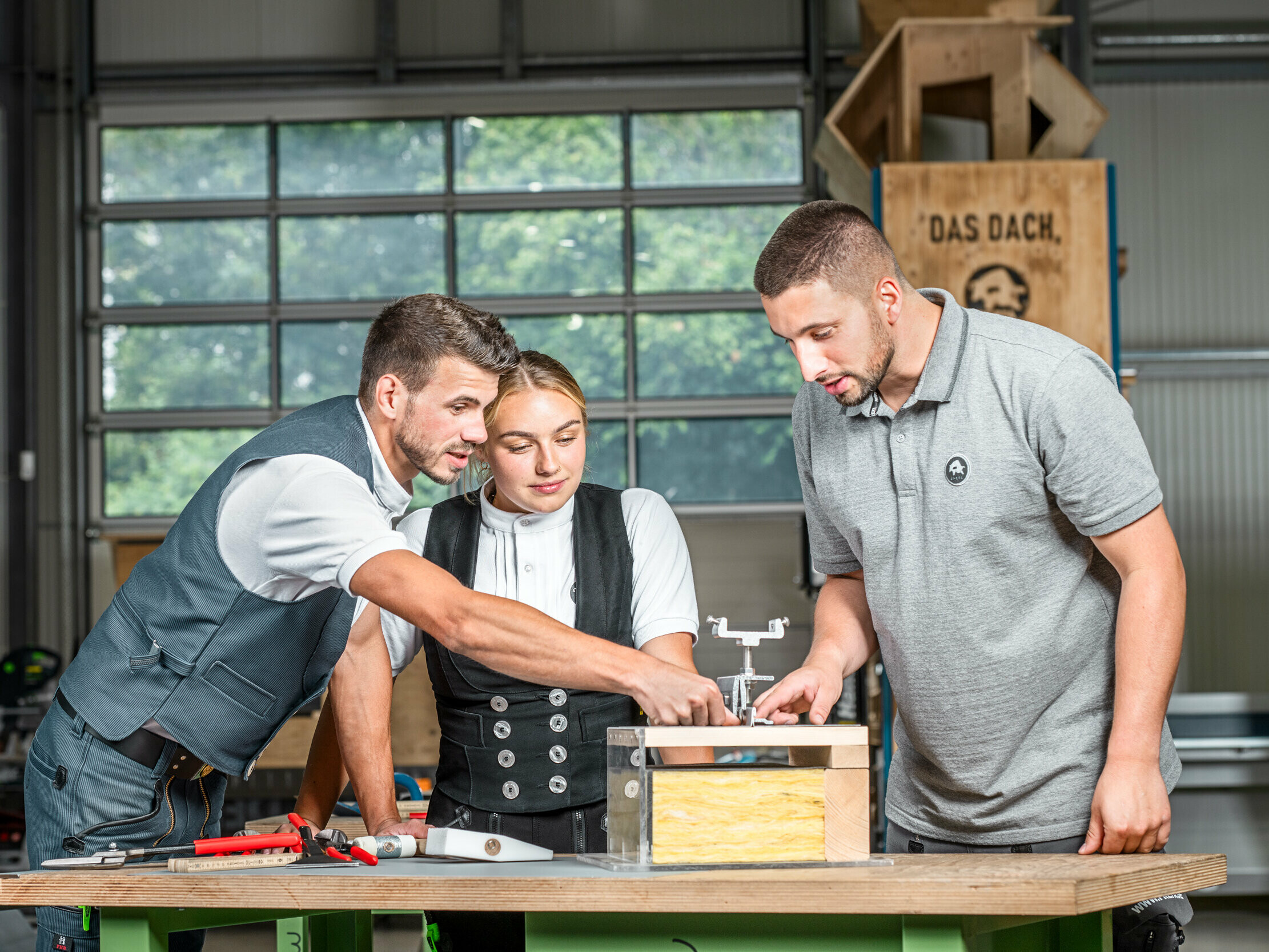 PREFA Anwendungstechniker Julian Eickhoff, Marvin Bürger von Bürger Falztechnik und Dachdeckermeisterin Fabienne Ellermeier schauen sich gemeinsam ein Lehrmodell in der PREFA Academy Rheinbach an. 