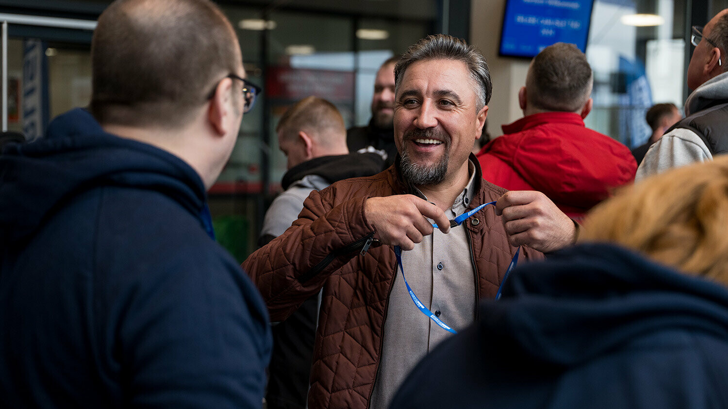 Eine lebendige Gesprächssituation während des Carlisle Branchenevents in der Mainz Arena. Ein Besucher steht im Mittelpunkt und lächelt, während er im Austausch mit anderen Teilnehmern ein Veranstaltungsband bzw. Badge in den Händen hält. Im Hintergrund sind weitere Gäste des Events erkennbar.