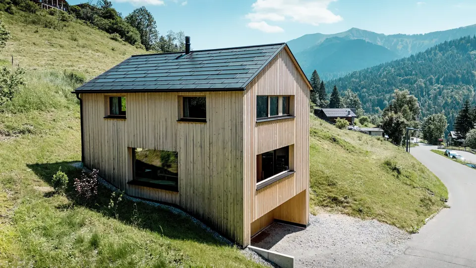 Hoch über dem Tal steht Haus Immi wie ein klarer Holzmonolith im sonnigen Berghang von Ebnit. Die reduzierte Weißtannenfassade und das schwarze PREFA Solardach setzen einen kraftvollen Kontrast zur weiten Alpenlandschaft. Integrierte Solardachplatten und Dachplatten R.16 in P.10 Schwarz liefern Solarstrom, trotzen alpinem Wetter und sichern ein langlebiges, harmonisches Dachbild.