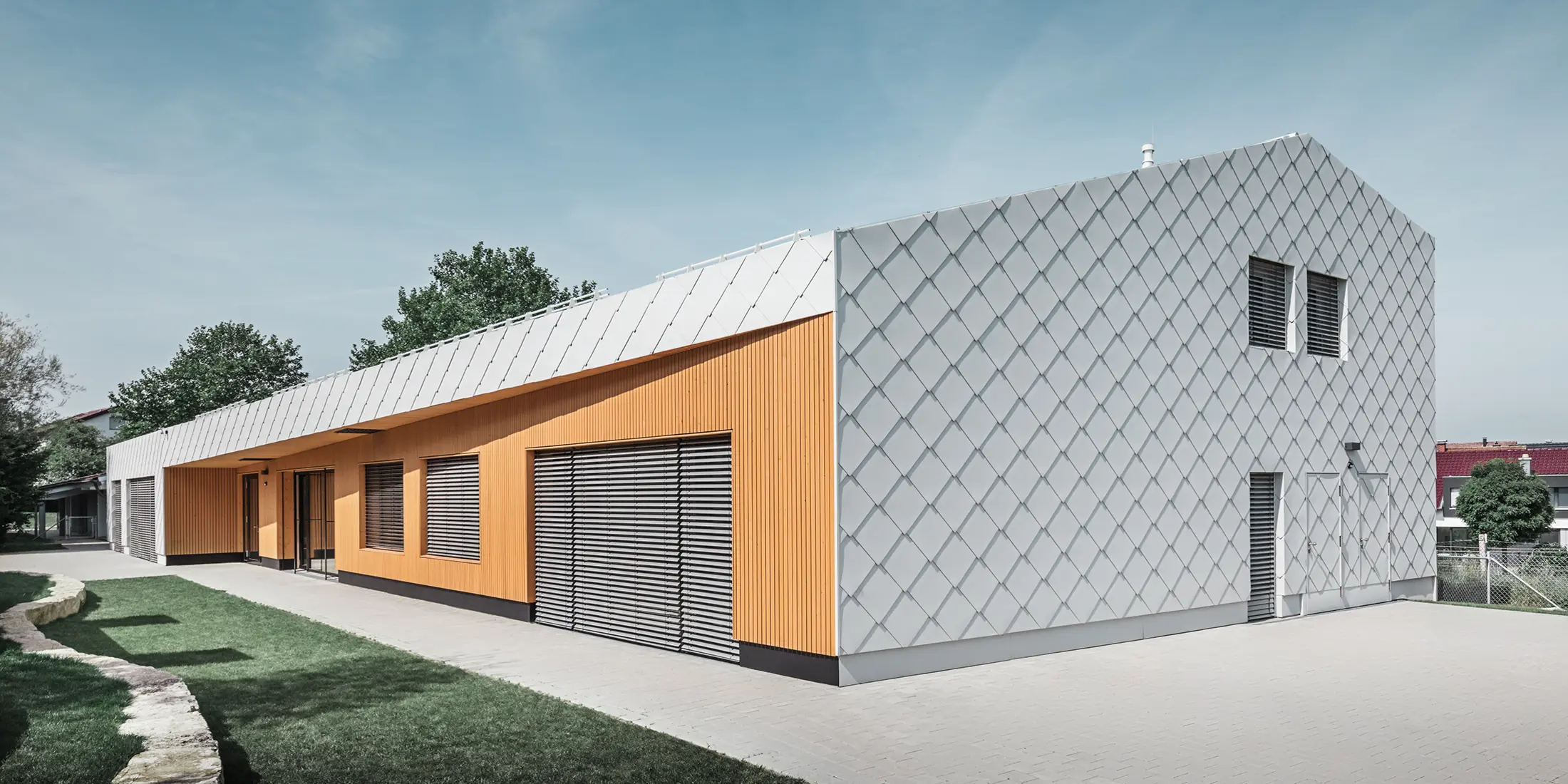 Modernes Kindergartengebäude mit Kombination aus silberner Aluminiumrautenfassade und Holzverkleidung in warmem Orangeton. Die Architektur vereint klare Linien mit funktionalen Elementen wie großen Fenstern und Sonnenschutzlamellen. Das Gebäude steht auf einem gepflasterten Gelände mit angrenzender Rasenfläche unter blauem Himmel.
