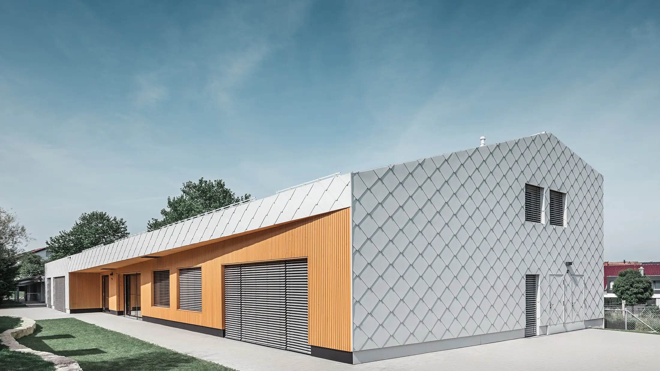 Modernes Kindergartengebäude mit Kombination aus silberner Aluminiumrautenfassade und Holzverkleidung in warmem Orangeton. Die Architektur vereint klare Linien mit funktionalen Elementen wie großen Fenstern und Sonnenschutzlamellen. Das Gebäude steht auf einem gepflasterten Gelände mit angrenzender Rasenfläche unter blauem Himmel.