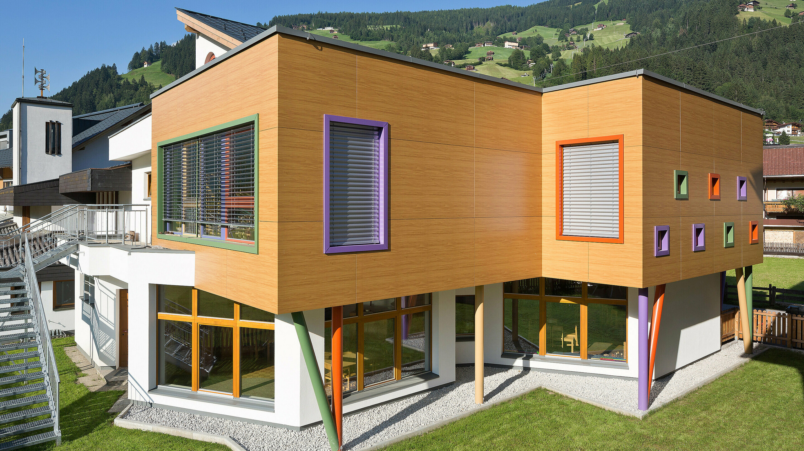 Modernes, zweigeschossiges Gebäude mit auffälliger Holzoptik-Fassade und farbenfrohen Akzenten. Fensterrahmen in Grün, Lila und Orange setzen lebendige Kontraste, während schräge, bunte Stützen die obere Etage tragen. Das Gebäude steht auf einer gepflegten Rasenfläche in einer alpinen Umgebung mit grünen Hügeln und Häusern im Hintergrund.