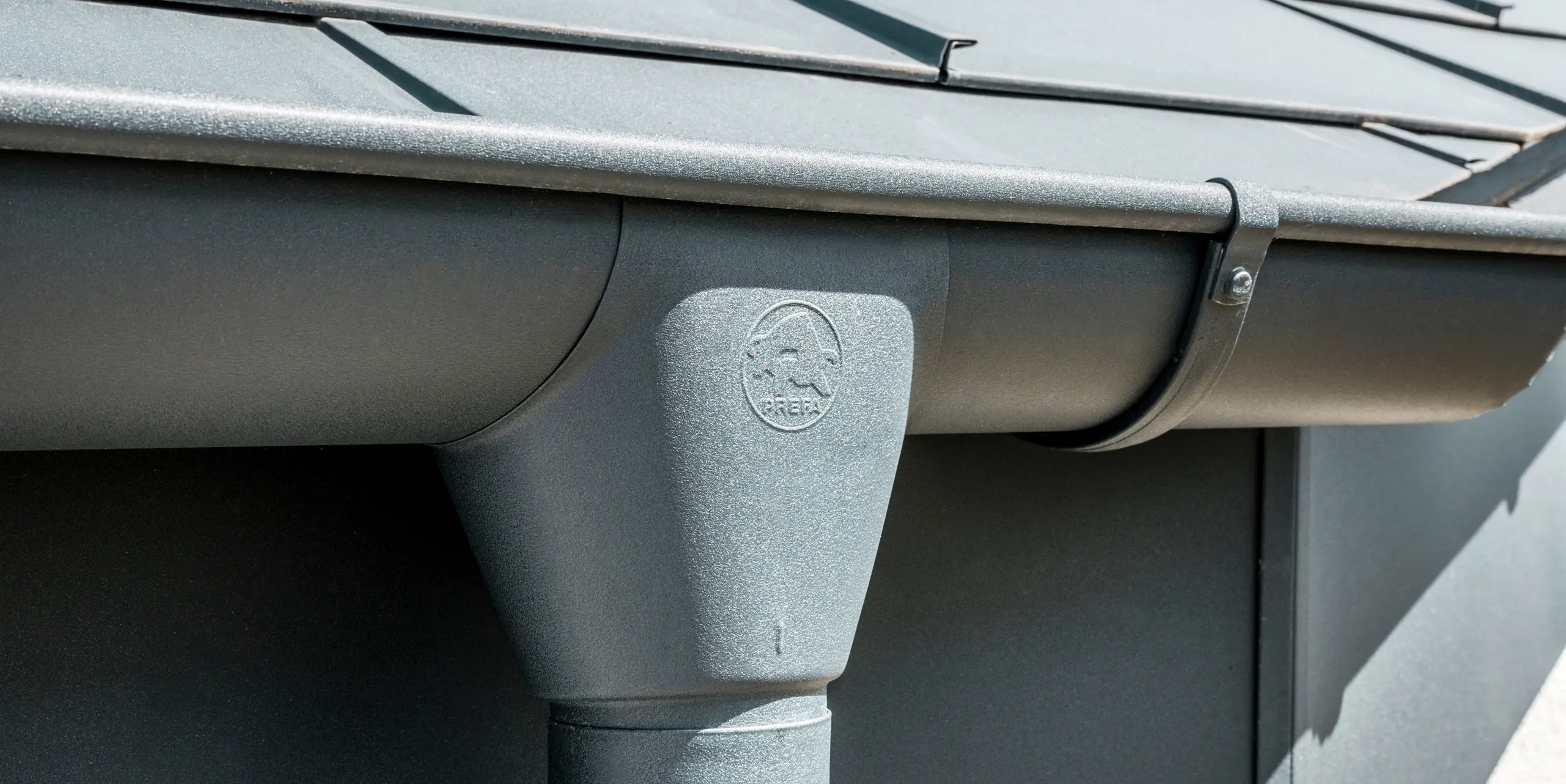 Nahaufnahme eines Hausdaches mit PREFA Dachplatte R.16 in Anthrazit sowie detailreicher Ansicht der farblich abgestimmten PREFA Dachrinne und Fallrohr aus Aluminium bei sonnigem Wetter.