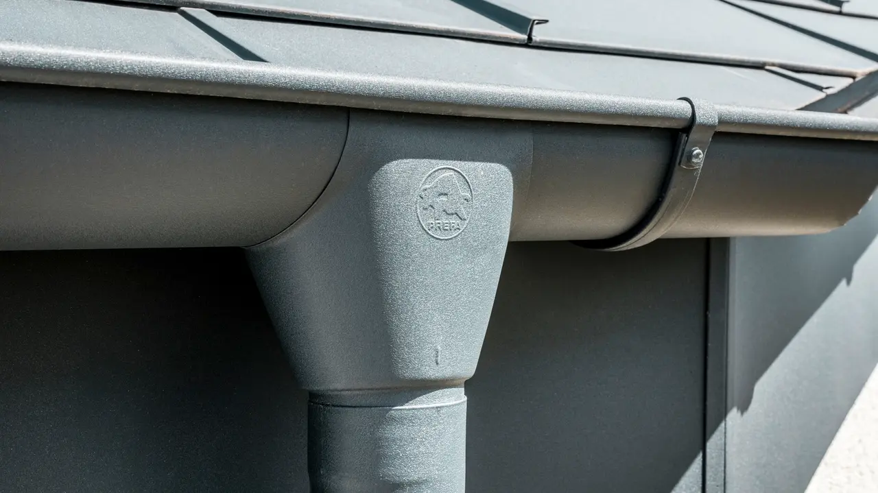 Nahaufnahme eines Hausdaches mit PREFA Dachplatte R.16 in Anthrazit sowie detailreicher Ansicht der farblich abgestimmten PREFA Dachrinne und Fallrohr aus Aluminium bei sonnigem Wetter.