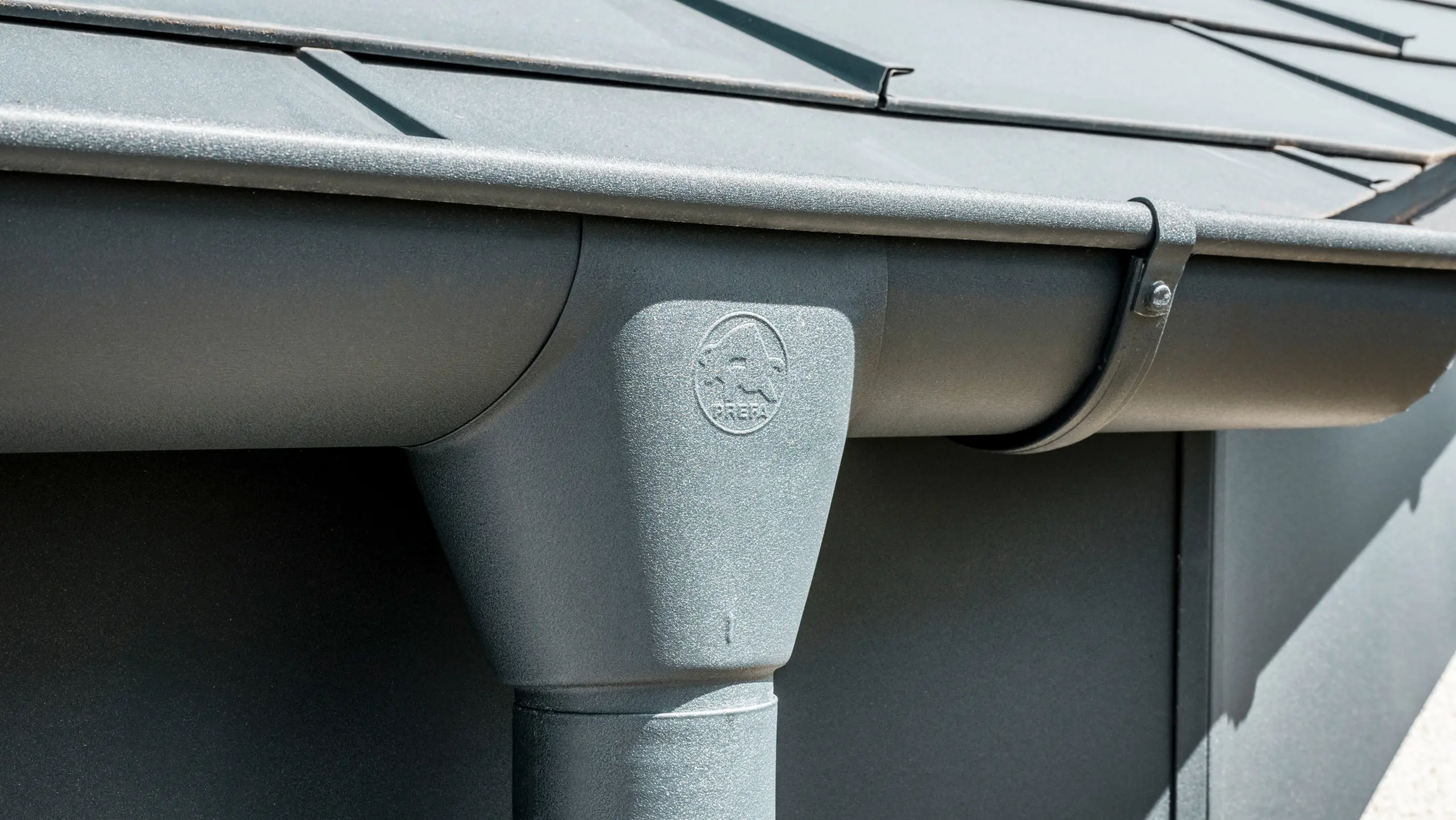 Nahaufnahme eines Hausdaches mit PREFA Dachplatte R.16 in Anthrazit sowie detailreicher Ansicht der farblich abgestimmten PREFA Dachrinne und Fallrohr aus Aluminium bei sonnigem Wetter.