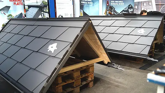 Zwei Übungsdächer mit dunkelgrauer Aluminium-Dacheindeckung in Schindeloptik stehen in der Werkhalle der PREFA Academy auf Holzpaletten. Die Modelle dienen dem Training im Klempnerhandwerk. Im Hintergrund sind Werbebanner mit dem PREFA-Logo und einer der teilnehmenden Meisterschüler zu sehen.