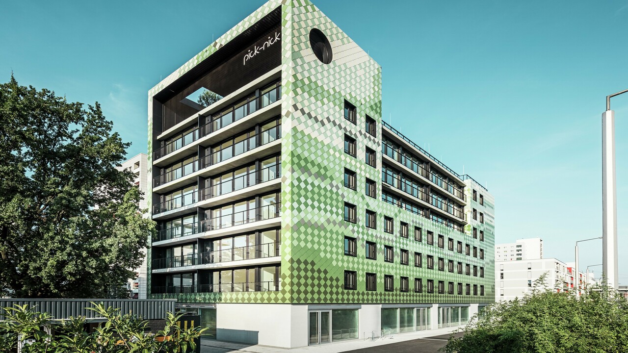 Das Wohn- und Geschäftshaus „Pick-Nick“ in Dresden besticht durch seine moderne PREFA-Aluminiumfassade in verschiedenen Grün- und Beigetönen. Die markante Fassade vereint klare Linien mit einem dynamischen Farbspiel und wird durch große Glasflächen zusätzlich betont. Architektonische Akzente setzen der Schriftzug „Pick-Nick“ an der oberen Gebäudekante und der begrünte Dachgarten.