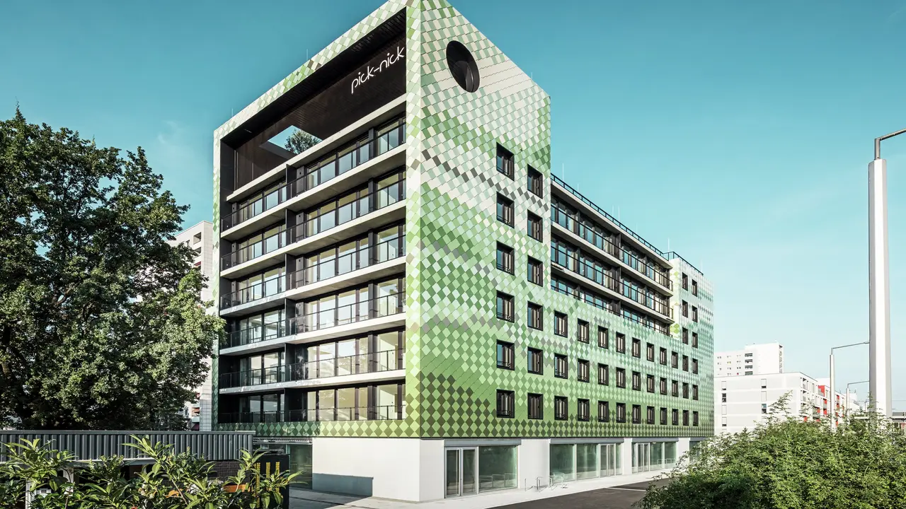 Das Wohn- und Geschäftshaus „Pick-Nick“ in Dresden besticht durch seine moderne PREFA-Aluminiumfassade in verschiedenen Grün- und Beigetönen. Die markante Fassade vereint klare Linien mit einem dynamischen Farbspiel und wird durch große Glasflächen zusätzlich betont. Architektonische Akzente setzen der Schriftzug „Pick-Nick“ an der oberen Gebäudekante und der begrünte Dachgarten.