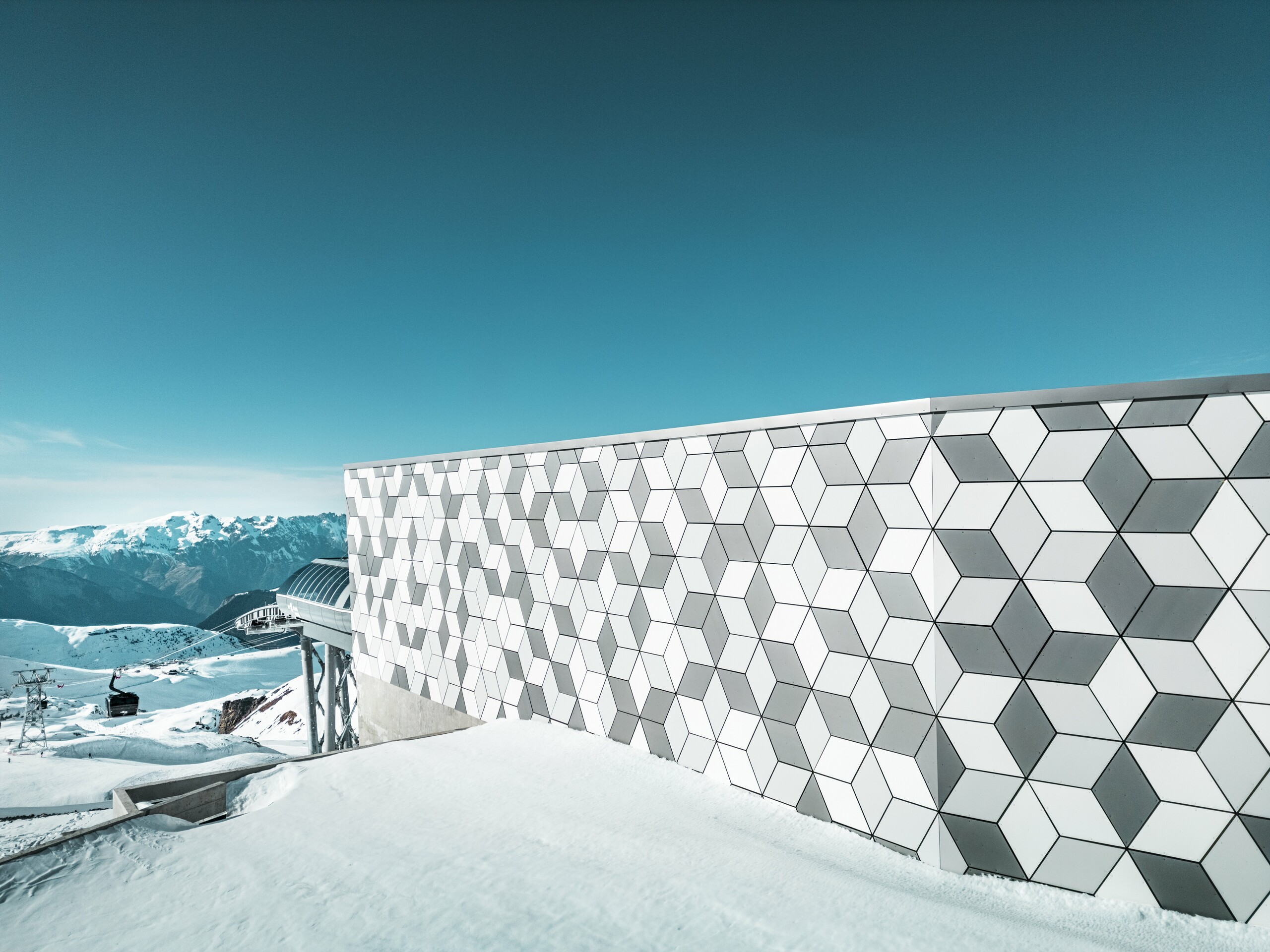 Aufnahme der Aluminiumfassade der Seilbahnstation in Les Deux Alpes: Die markante Fassade aus PREFABOND Aluminiumverbundplatten beeindruckt mit ihrer auffälligen, geometrischen Rautenmusteranordnung. Die harmonische Kombination aus Weiß und Grau erzeugt einen modernen Look, der sich nahtlos in die majestätische Berglandschaft einfügt. Hinter der Fassade sind der strahlend blaue Himmel und das majestätische Bergpanorama zu sehen.