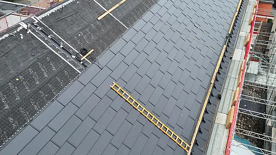 Vogelperspektive auf das teilweise neu sanierte Dach der St.-Karl-Borromäus-Kirche, das mit großen PREFA Solardachplatten ausgestattet ist. Die Platten sind gleichmäßig auf dem gesamten Dach verlegt und haben eine dunkle, glänzende Oberfläche. Die Architektur der Kirche ist im Hintergrund sichtbar, einschließlich der charakteristischen Dachform und des Turms. Die Solardachplatten sind präzise montiert und passen sich optisch gut in das denkmalgeschützte Gebäude ein.