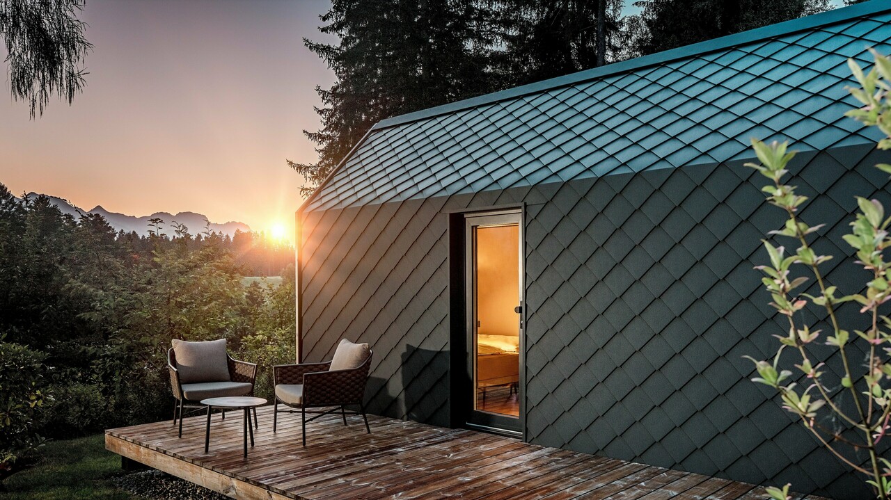 Ein minimalistisches Tiny House im Secret Koi Garden am Natterer See zeigt sich bei Sonnenuntergang mit vorgelagerter Holzterrasse und Blick in den Wald. Dach und Fassade sind mit kleinteiligen PREFA Dach‑ und Wandrauten aus Aluminium in P.10 Anthrazit ausgeführt, deren strukturierte Oberfläche das warme Licht eindrucksvoll reflektiert. Das langlebige, witterungsbeständige PREFA Aluminium schützt die Architektur zuverlässig und schafft einen markanten Kontrast zu Holz, Natur und sanfter Beleuchtung.
