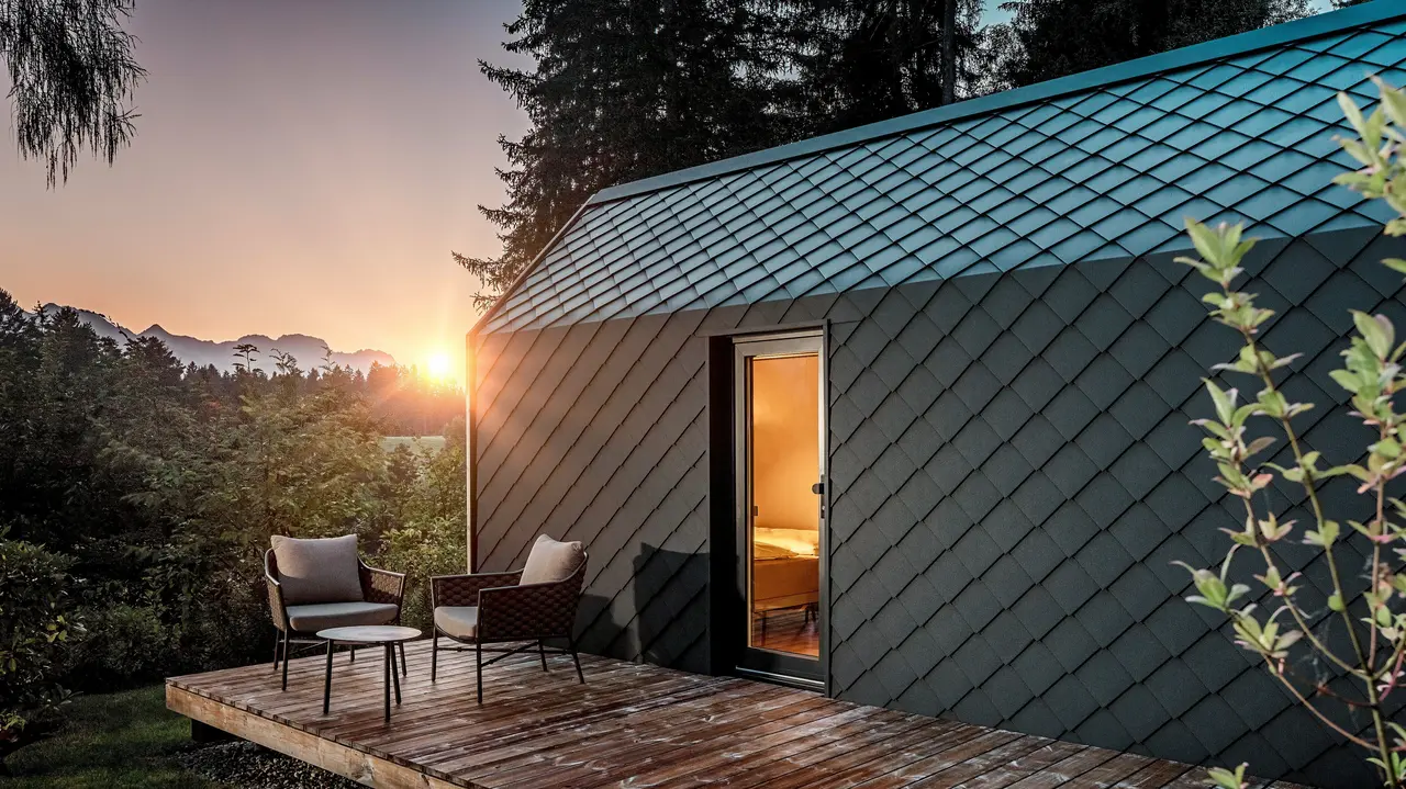 Ein minimalistisches Tiny House im Secret Koi Garden am Natterer See zeigt sich bei Sonnenuntergang mit vorgelagerter Holzterrasse und Blick in den Wald. Dach und Fassade sind mit kleinteiligen PREFA Dach‑ und Wandrauten aus Aluminium in P.10 Anthrazit ausgeführt, deren strukturierte Oberfläche das warme Licht eindrucksvoll reflektiert. Das langlebige, witterungsbeständige PREFA Aluminium schützt die Architektur zuverlässig und schafft einen markanten Kontrast zu Holz, Natur und sanfter Beleuchtung.