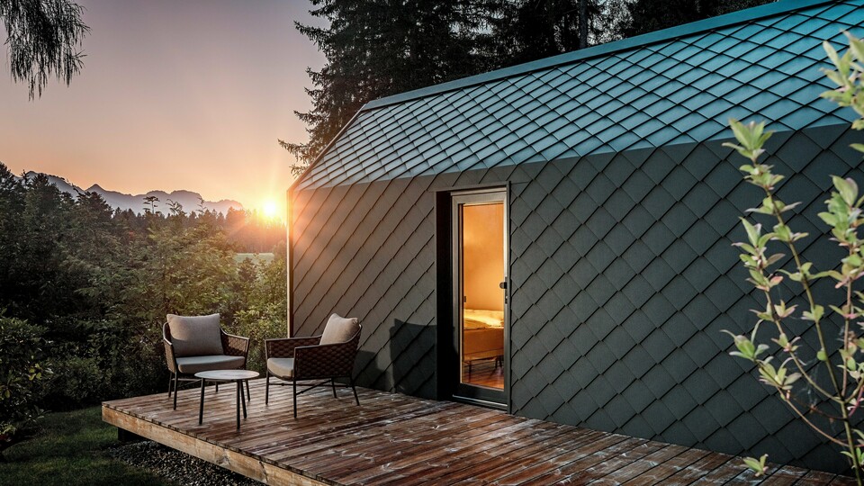 Ein minimalistisches Tiny House im Secret Koi Garden am Natterer See zeigt sich bei Sonnenuntergang mit vorgelagerter Holzterrasse und Blick in den Wald. Dach und Fassade sind mit kleinteiligen PREFA Dach‑ und Wandrauten aus Aluminium in P.10 Anthrazit ausgeführt, deren strukturierte Oberfläche das warme Licht eindrucksvoll reflektiert. Das langlebige, witterungsbeständige PREFA Aluminium schützt die Architektur zuverlässig und schafft einen markanten Kontrast zu Holz, Natur und sanfter Beleuchtung.