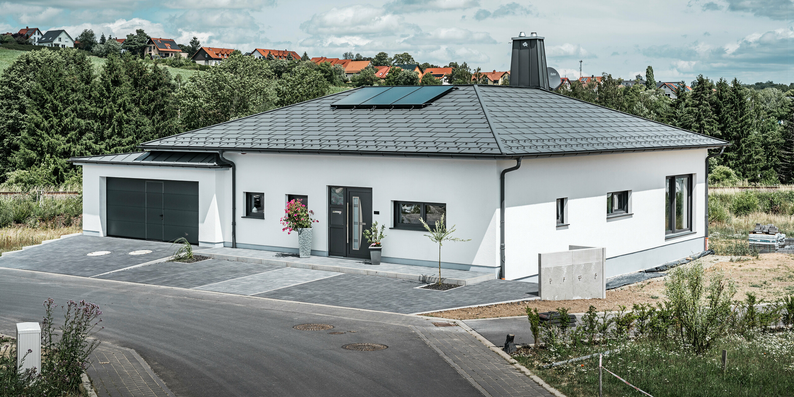 Elegantes Einfamilienhaus in Ilmenau, Thüringen, mit einem hochwertigen PREFA Dach in der Farbe P.10 Dunkelgrau, ausgestattet mit der robusten Dachplatte R.16. Die Garage harmoniert in Farbe und Stil durch die Verwendung von PREFALZ. Der Blickfang des Anwesens ist das funktional gestaltete Atrium und der mit dem PREFA Stier verzierte Schornstein, welcher das moderne Design und die innovative Dachtechnologie von PREFA unterstreichen. Die Solarpaneele fügen sich nahtlos in das nachhaltige Konzept des Hauses ein. Abgerundet wird das durchdachte Gesamtkonzept des Bungalows durch das PREFA Dachentwässerungssytem, welches ebenfalls in Dunkelgrau gehalten ist.