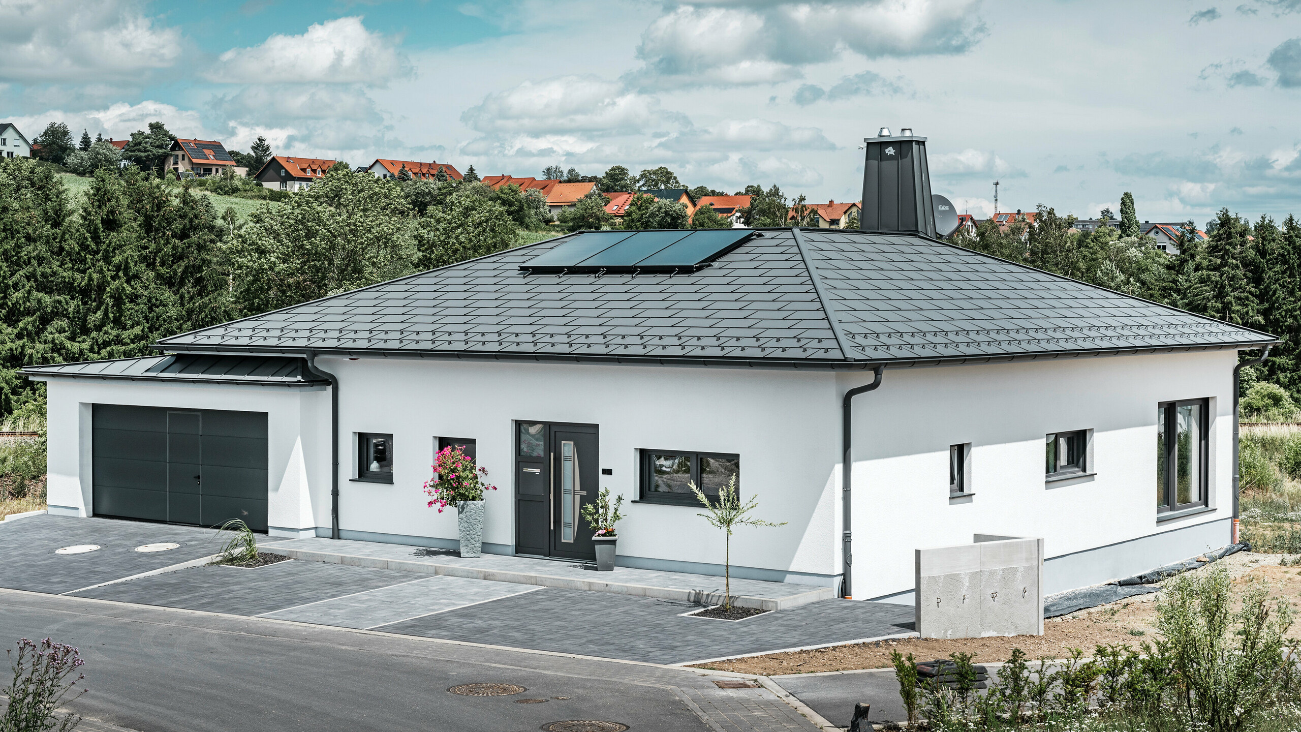 Elegantes Einfamilienhaus in Ilmenau, Thüringen, mit einem hochwertigen PREFA Dach in der Farbe P.10 Dunkelgrau, ausgestattet mit der robusten Dachplatte R.16. Die Garage harmoniert in Farbe und Stil durch die Verwendung von PREFALZ. Der Blickfang des Anwesens ist das funktional gestaltete Atrium und der mit dem PREFA Stier verzierte Schornstein, welcher das moderne Design und die innovative Dachtechnologie von PREFA unterstreichen. Die Solarpaneele fügen sich nahtlos in das nachhaltige Konzept des Hauses ein. Abgerundet wird das durchdachte Gesamtkonzept des Bungalows durch das PREFA Dachentwässerungssytem, welches ebenfalls in Dunkelgrau gehalten ist.