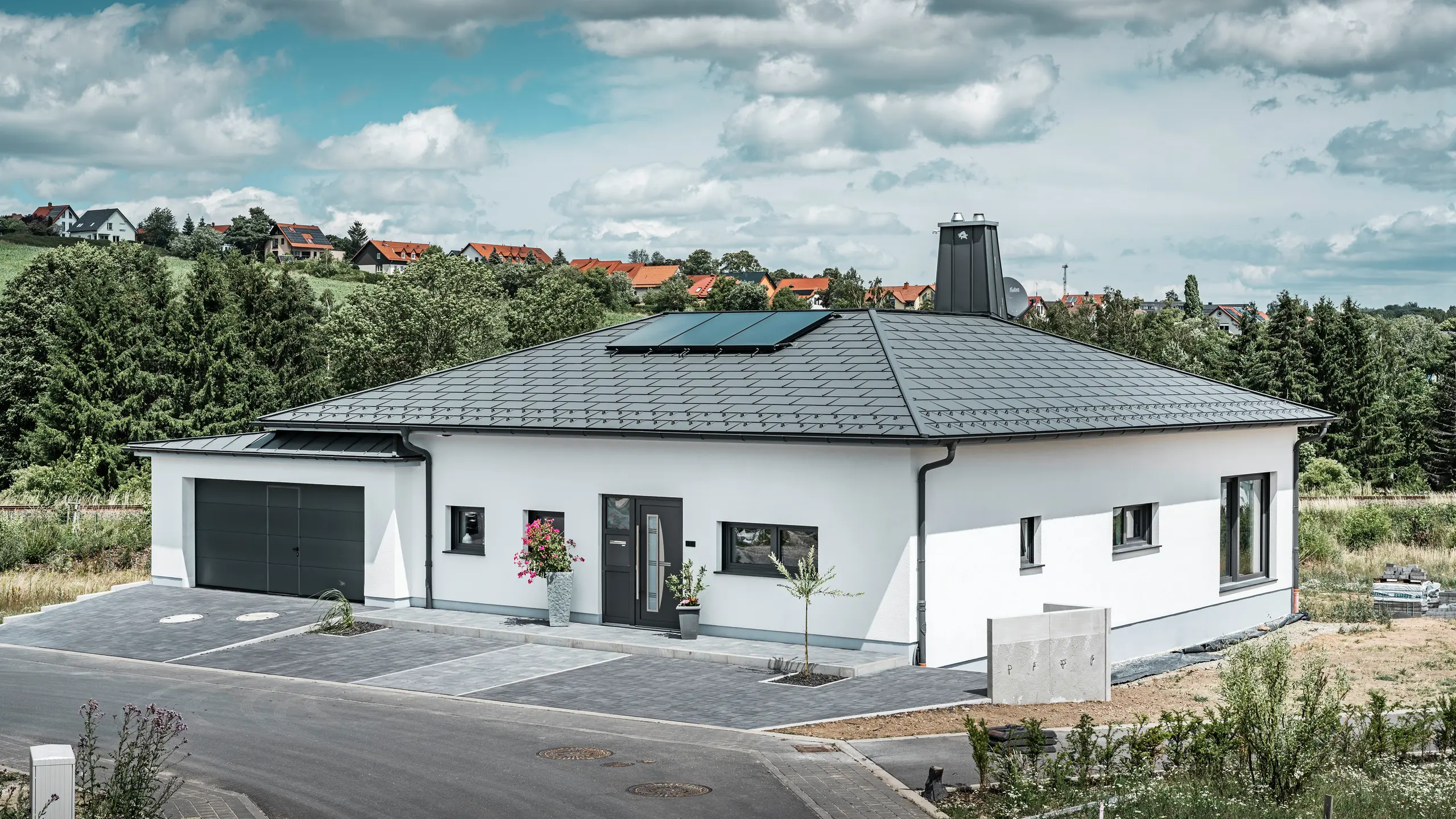 Elegantes Einfamilienhaus in Ilmenau, Thüringen, mit einem hochwertigen PREFA Dach in der Farbe P.10 Dunkelgrau, ausgestattet mit der robusten Dachplatte R.16. Die Garage harmoniert in Farbe und Stil durch die Verwendung von PREFALZ. Der Blickfang des Anwesens ist das funktional gestaltete Atrium und der mit dem PREFA Stier verzierte Schornstein, welcher das moderne Design und die innovative Dachtechnologie von PREFA unterstreichen. Die Solarpaneele fügen sich nahtlos in das nachhaltige Konzept des Hauses ein. Abgerundet wird das durchdachte Gesamtkonzept des Bungalows durch das PREFA Dachentwässerungssytem, welches ebenfalls in Dunkelgrau gehalten ist.