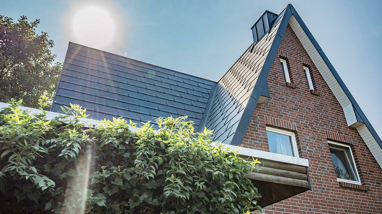 Satteldach mit der schlichten PREFA Alu-Dachplatte R.16 in Anthrazit eingedeckt. Die Fassade ist eine rustikale Klinkerfassade, hinter dem Haus geht gerade die Sonne auf.