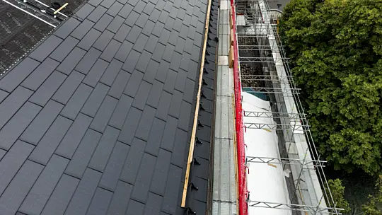 Vogelperspektive auf die bereits verlegten PREFA Solardachplatten auf dem Dach der St.-Karl-Borromäus-Kirche in Nürnberg. Die Platten sind gleichmäßig auf dem Dach installiert und haben eine schwarze, glänzende Oberfläche. Die Kirche ist im Hintergrund des Bildes sichtbar und zeigt ihre traditionelle Architektur mit einem steilen Dach und einem spitzen Turm. Die Solardachplatten sind in klaren Reihen angeordnet, die sich über die gesamte Dachfläche erstrecken.