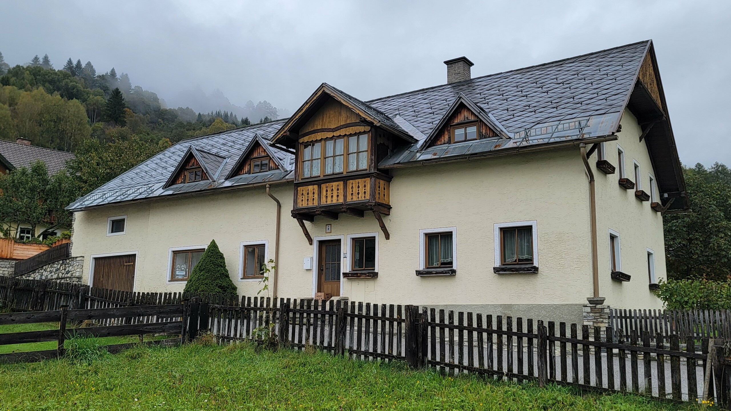 Ein traditionelles Einfamilienhaus im Alpenraum bei Regenwetter mit einem stark verwitterten Eternit-Dach. Die ehemals glatten Dachflächen wirken unregelmäßig und stumpf. Die Rinne ist veraltet und das gesamte Erscheinungsbild ist matt und wenig einladend. Der dunkle Himmel verstärkt die triste Wirkung der Fassade. Diese Ausgangssituation verdeutlicht die Notwendigkeit einer hochwertigen Dachsanierung. Die Szene bereitet den Kontrast zur neuen PREFA-Lösung vor und betont die gestalterischen und funktionalen Schwächen der alten Dacheindeckung.