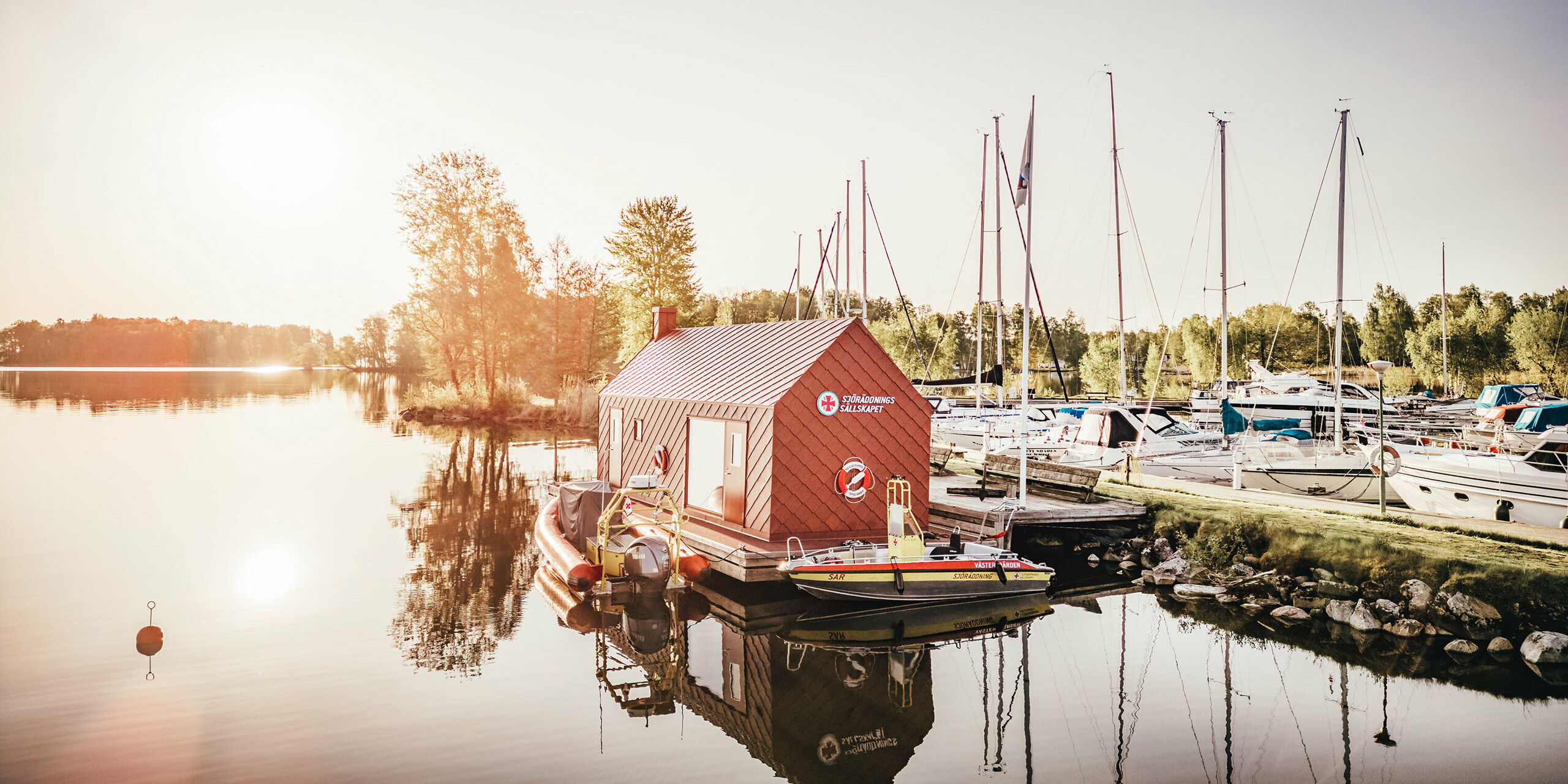 Frühe Morgenstunden am Hjälmaren See in Schweden, wo das schwimmende Seenotrettungsstationshaus mit einer oxydroten Aluminiumhülle aus PREFA Dach- und Wandrauten 29×29 ruhig auf dem Wasser liegt. Die aufgehende Sonne hinter dem Gebäude taucht die Szene in ein sanftes Licht, das sich zusammen mit dem Stationshaus und den Rettungsbooten im stillen Wasser spiegelt. Im Hintergrund reihen sich die Masten der im Hafen vertäuten Segelboote auf und vervollständigen das Bild eines friedlichen Rückzugsortes, der zugleich ein lebensrettender Stützpunkt ist.