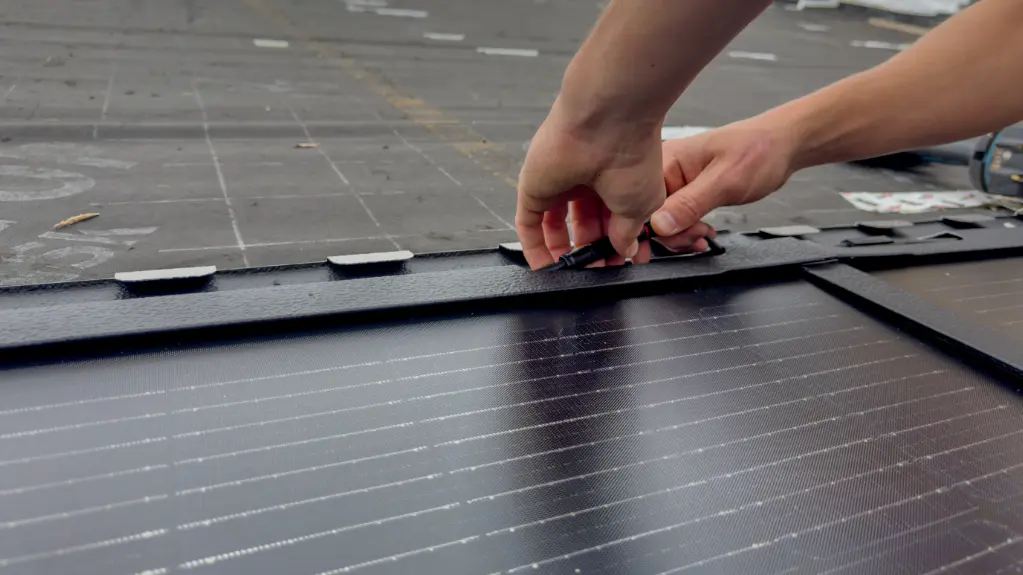 Nahaufnahme der Installation einer großen PREFA Solardachplatte auf dem Dach der St. Karl Borromäus Kirche in Nürnberg (Bayern). Die glänzenden, rechteckigen Platten sind sauber nebeneinander aufgereiht und haben eine schwarze Oberfläche. Die Hände des Handwerkers sind im rechten Teil des Bildes zu sehen. Er schließt die Kabel der Solardachplatte an. Der Hintergrund zeigt das bereits teilweise verlegte und vorbereitete Dach. Die Szene findet unter freiem Himmel statt, was auf die Außenmontage der Solarmodule hinweist.