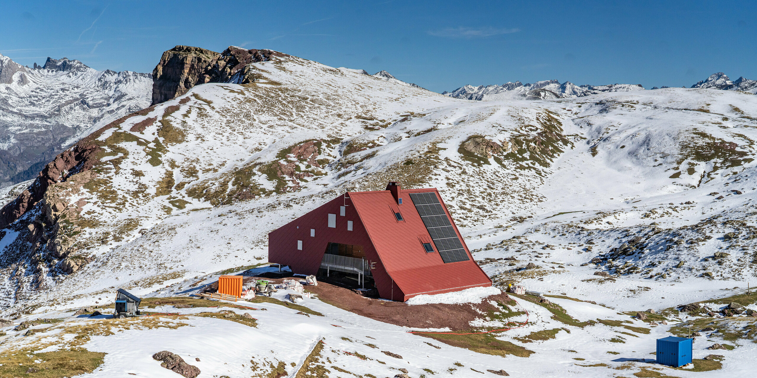 Vogelperspektive auf Schutzhütte Arlet im Pyrenäen-Nationalpark mit schneebedeckter Berglandschaft rund um das oxydrote Gebäude mit einem Dach und einer Fassade aus Aluminium