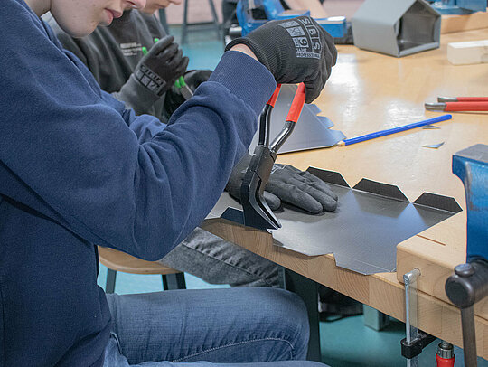 Ein Schüler verwendet eine Zange, um das Aluminiumblech für das Vogelhaus zu bearbeiten. Der Schüler trägt Schutzhandschuhe und konzentriert sich auf die präzise Arbeit. Im Hintergrund ist ein weiterer Schüler zu sehen, der ebenfalls an einem ähnlichen Blech arbeitet. Auf dem Tisch sind verschiedene Werkzeuge und Materialien sichtbar, die für den Bau der Vogelhäuser verwendet werden.