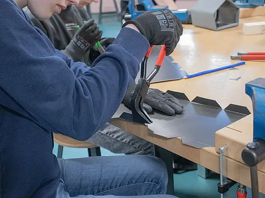 Ein Schüler verwendet eine Zange, um das Aluminiumblech für das Vogelhaus zu bearbeiten. Der Schüler trägt Schutzhandschuhe und konzentriert sich auf die präzise Arbeit. Im Hintergrund ist ein weiterer Schüler zu sehen, der ebenfalls an einem ähnlichen Blech arbeitet. Auf dem Tisch sind verschiedene Werkzeuge und Materialien sichtbar, die für den Bau der Vogelhäuser verwendet werden.