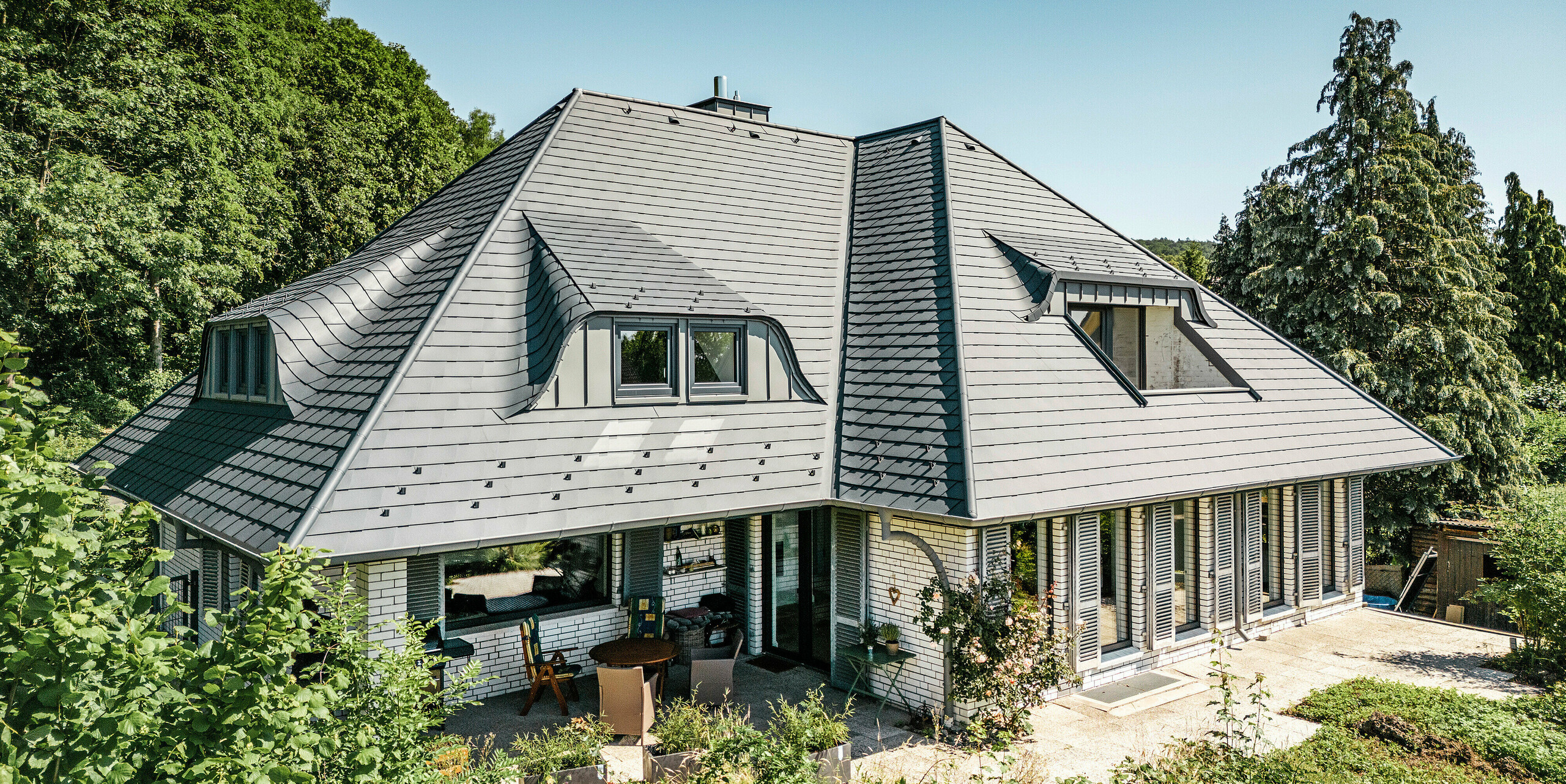 Modernes Einfamilienhaus in Einbeck, Deutschland, mit PREFA Dachschindeln in P.10 Anthrazit. Die langlebigen PREFA Dachschindeln aus Aluminium sind für ihre Qualität und Langlebigkeit bekannt. Die Dachgestaltung des Gebäudes betont die individuelle Architektur mit Hechtgauben, die durch die Flexibilität der PREFA Dachschindeln hervorgehoben wird. Die Dachentwässerung in Form des PREFA Ablaufrohrs bzw. der Hängerinne ist ebenfalls in Anthrazit gehalten. Rund um das Einfamilienhaus ist ein großzügiger und gepflegter Garten sowie eine Terrasse zu sehen. Hinter dem Haus liegt ein Wald, der - gemeinsam mit dem blauen Himmel - eine natürliche Atmosphäre schafft. 
