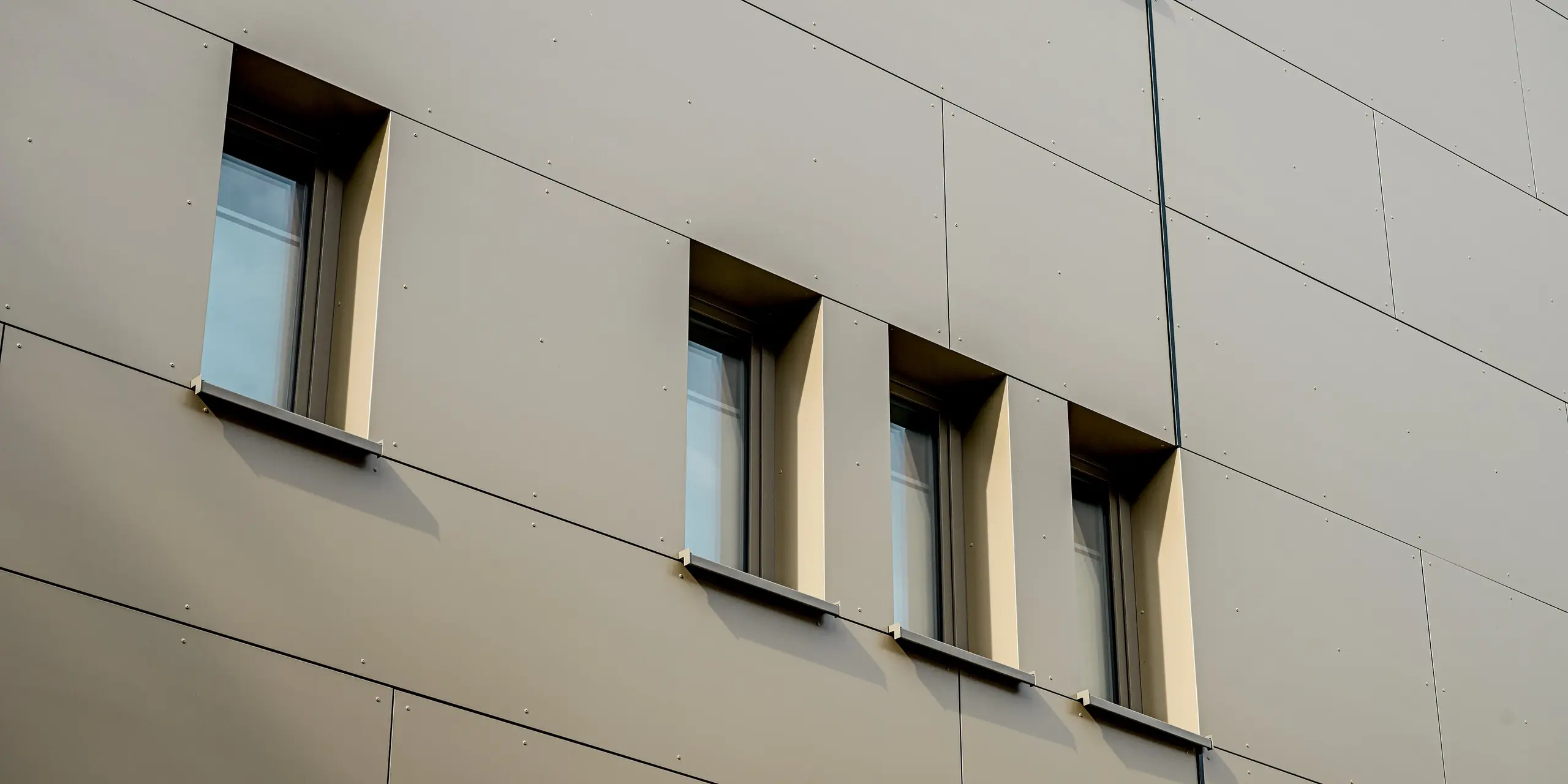 Das Bild zeigt eine Detailaufnahme der Fassade des Festo-Gebäudes in Langenhagen. Hier kommen die eleganten PREFABOND Aluminium Verbundplatten zum Einsatz, die in einem sanften Bronze gehalten sind und der Wand eine ruhige, moderne Ästhetik verleihen. Die Fenster sind in speziell geformte Aussparungen eingefasst, was den minimalistischen Charakter des Gebäudes unterstreicht. Die präzise Anordnung der Fenster und die feine Linienführung betonen das klare Design. Dank der hochfesten und witterungsbeständigen Materialien bleibt die Fassade über Jahre hinweg ästhetisch und funktional, perfekt für langlebige Architektur.