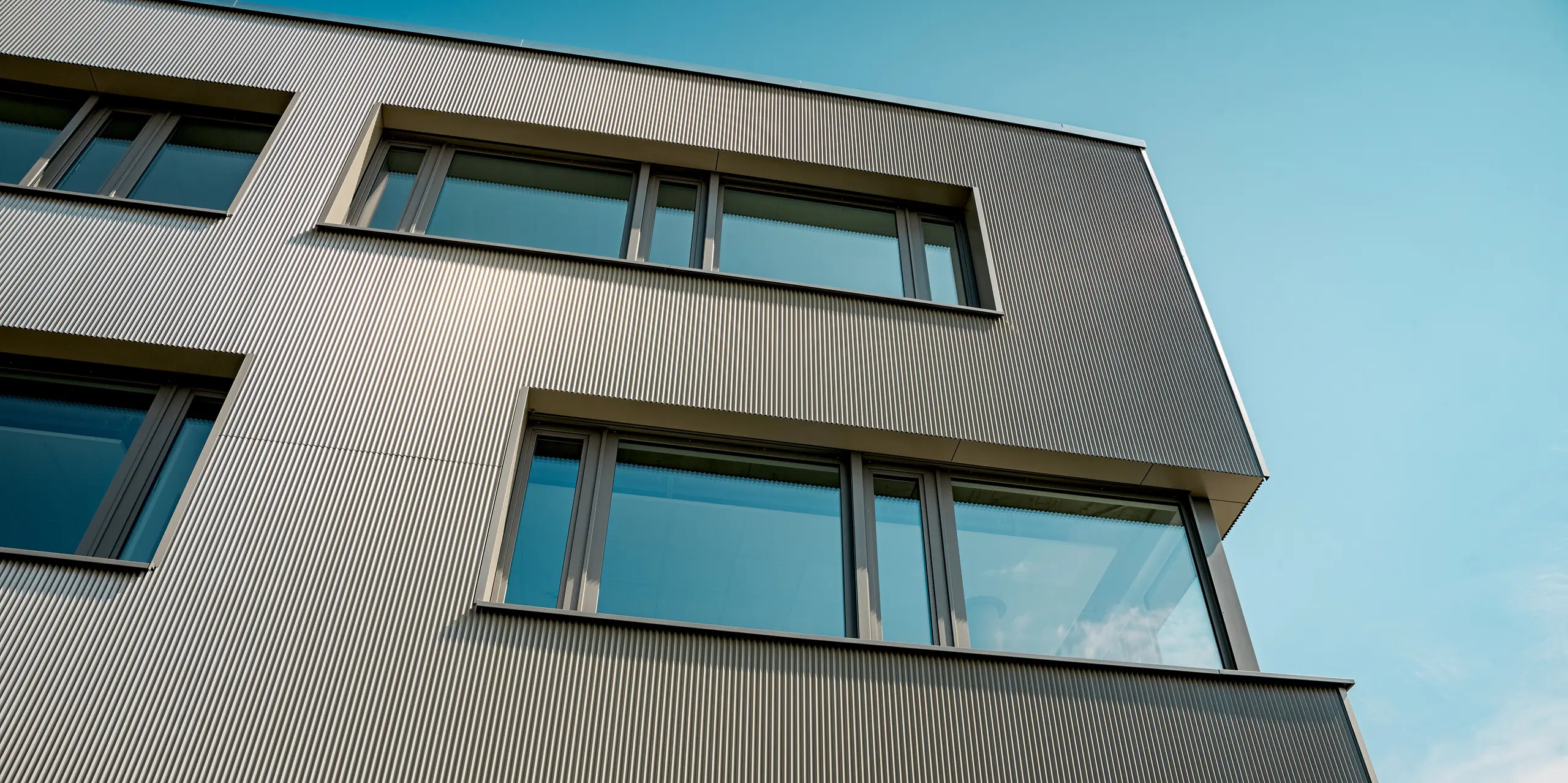 Das Bild zeigt eine Detailaufnahme der Aluminiumfassade des Festo-Gebäudes in Langenhagen, die mit PREFA Strangpressprofilen in Zackenform ausgestattet ist. Die vertikale Struktur der Profile verleiht der Fassade eine markante, texturierte Optik, die das Gebäude sowohl modern als auch dynamisch erscheinen lässt. Besonders beeindruckend ist die klare Linienführung und die präzise Integration der Fenster, die eine harmonische Balance zwischen Architektur und Funktionalität bieten. Der Einsatz der robusten und langlebigen PREFA Produkte sorgt für eine ästhetische und witterungsbeständige Fassade, die den hohen Anforderungen an Design und Nachhaltigkeit gerecht wird.