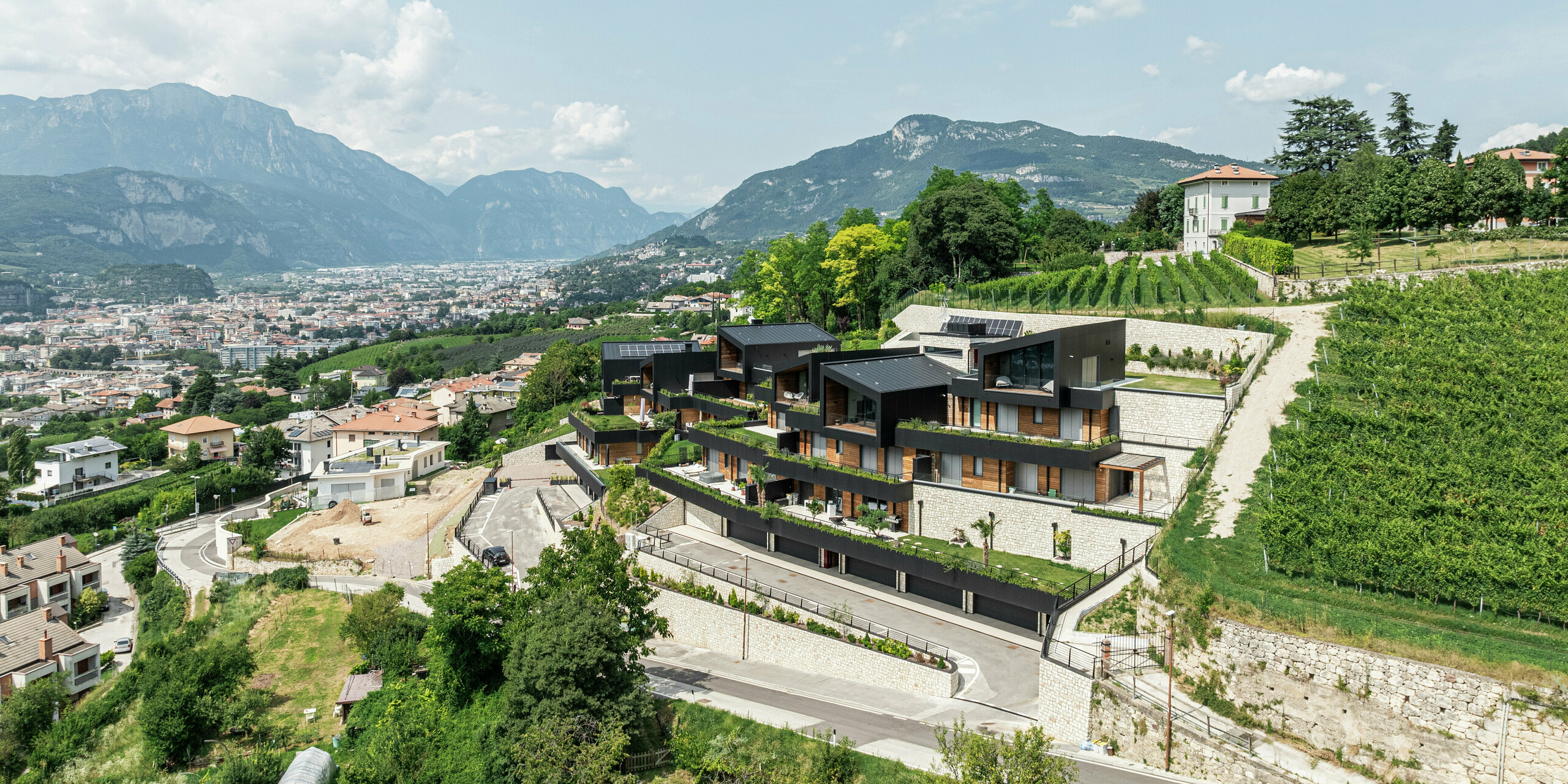 Spektakuläre Hanglage mit Blick auf Trient: Die exklusive Wohnanlage fügt sich perfekt in die umliegenden Weinberge und die Hügellandschaft ein. Die moderne Architektur verbindet Naturstein, Holz und die langlebige PREFA Aluminiumfassade in P.10 Schwarz zu einer eleganten und witterungsbeständigen Einheit. Das PREFALZ Dach in P.10 Schwarz sorgt für maximalen Schutz und Langlebigkeit. Großzügige Terrassen und Begrünungen unterstreichen das nachhaltige Wohnkonzept und schaffen ein harmonisches Zusammenspiel von Natur und moderner Bauweise.