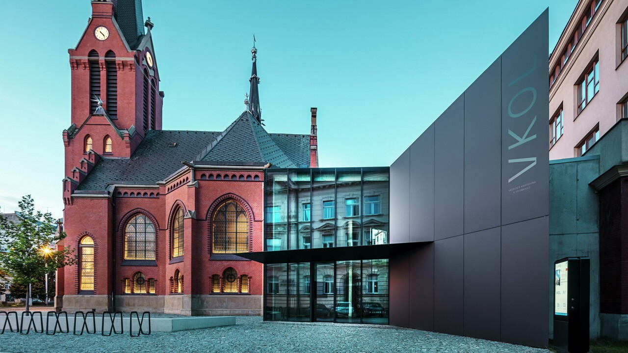 Das Bild zeigt das imposante Zusammenspiel zwischen der historischen Roten Kirche und dem modernen Neubau in Olmütz. Die Kirche mit ihren charakteristischen gotischen Fenstern und dem markanten Turm bildet das historische Rückgrat des Ensembles, während der moderne Anbau mit seiner eleganten, schwarzen PREFABOND Aluminium-Verbundplatten-Fassade eine zeitgemäße, architektonische Antwort darstellt. Große Glasflächen lassen den Blick in das Innere des Neubaus zu, wo die Umgebung durch Reflexionen in den Fenstern eine interessante Wechselwirkung zwischen Alt und Neu widerspiegelt. Die präzise Verarbeitung der PREFABOND-Platten betont die moderne Ästhetik, während die harmonische Integration in das historische Umfeld sowohl funktional als auch visuell beeindruckt.