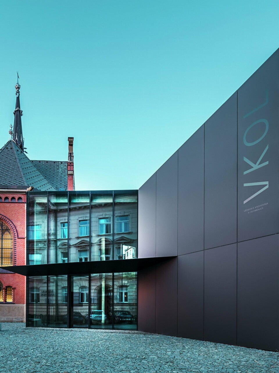 Das Bild zeigt das imposante Zusammenspiel zwischen der historischen Roten Kirche und dem modernen Neubau in Olmütz. Die Kirche mit ihren charakteristischen gotischen Fenstern und dem markanten Turm bildet das historische Rückgrat des Ensembles, während der moderne Anbau mit seiner eleganten, schwarzen PREFABOND Aluminium-Verbundplatten-Fassade eine zeitgemäße, architektonische Antwort darstellt. Große Glasflächen lassen den Blick in das Innere des Neubaus zu, wo die Umgebung durch Reflexionen in den Fenstern eine interessante Wechselwirkung zwischen Alt und Neu widerspiegelt. Die präzise Verarbeitung der PREFABOND-Platten betont die moderne Ästhetik, während die harmonische Integration in das historische Umfeld sowohl funktional als auch visuell beeindruckt.