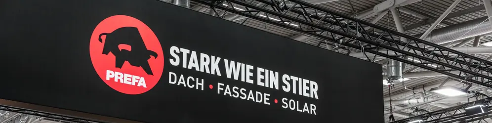 Großes PREFA Logo mit dem Slogan „Stark wie ein Stier – Dach, Fassade, Solar“ auf einer hängenden Werbetafel am Messestand auf der BAU Messe. Im Hintergrund sind Visualisierungen moderner Architekturprojekte mit PREFA Fassadenlösungen sowie Teile anderer Messestände zu sehen.