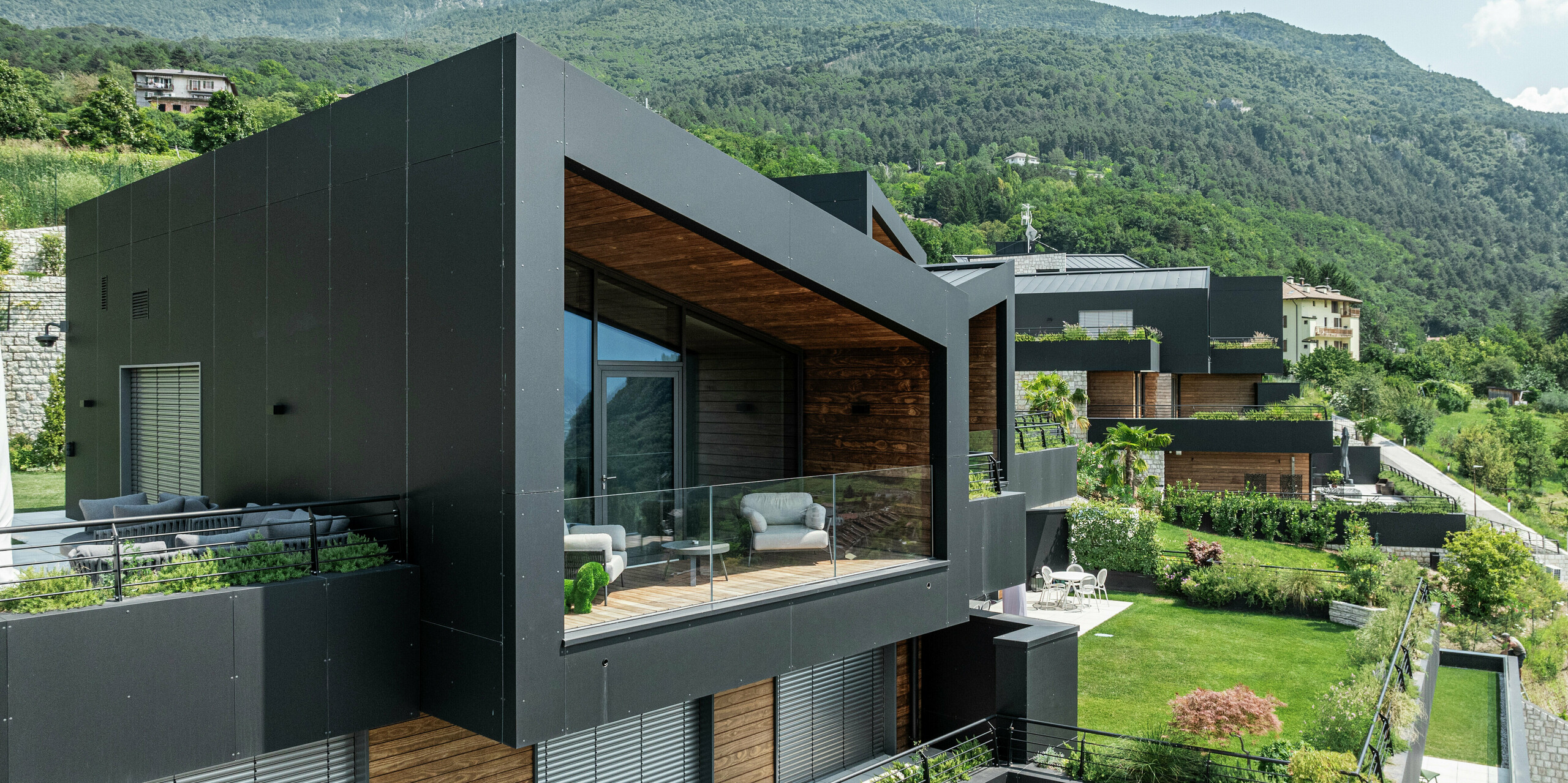 Zeitgemäße Architektur inmitten der Natur: Die großzügige Glasfront dieses Wohnhauses eröffnet einen atemberaubenden Blick auf die grüne Hügellandschaft. Die Fassade aus PREFA Aluminium Verbundplatten in P.10 Schwarz sorgt für eine elegante, witterungsbeständige Gebäudehülle, die im Kontrast zum warmen Holz steht. Ergänzt wird das moderne Design durch eine überdachte Terrasse mit Glasgeländer, die Innen- und Außenbereich nahtlos miteinander verbindet. Dank der langlebigen und pflegeleichten PREFA Materialien bleibt die Optik dauerhaft ansprechend - perfekt für nachhaltiges Wohnen mit Stil.