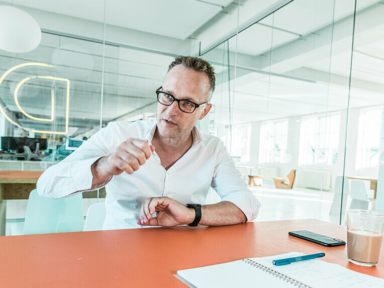 Architekt Noel Wibrand sitzt mit dem Gesicht zur Kamera an einem Schreibtisch und erklärt etwas