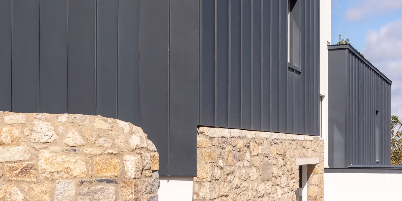 Die Aufnahme zeigt die moderne Fassade eines Reihenhauses in Watson Place mit klaren Linien und einer markanten Kombination aus Natursteinmauer und dunkel anmutender PREFALZ Verkleidung in P.10 Anthrazit. Die stehenden Falze des Aluminiumprofils setzen vertikale Akzente und verleihen dem Gebäude eine elegante Struktur. Das witterungsbeständige PREFA Aluminium schützt dauerhaft vor Feuchtigkeit und Wind, während die leichte Bauweise flexible Gestaltung ermöglicht. Das Zusammenspiel aus robustem Naturstein und hochwertigem Aluminium schafft einen zugleich zeitlosen und dynamischen Gesamteindruck.